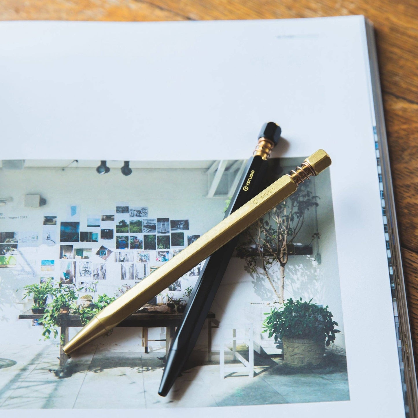 Zwei ENTREPÔT - YSTUDIO - Klassischer Revolve Kugelschreiber Feder (Messing) in Schwarz und Gold, mit Federkappenstruktur, liegen gekreuzt auf einem Magazin, das zu einem hellen, dekorierten Zimmerfoto mit Pflanzen aufgeschlagen ist, das den langlebigen Stil zeigt.