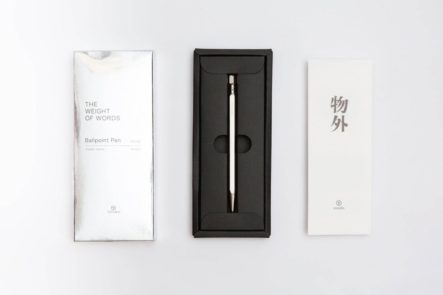 Ein silberglänzender ENTREPÔT - YSTUDIO Klassischer Drehkugelschreiber Feder ruht in einer schwarzen Schachtel mit zwei weißen Kärtchen - auf dem einen steht "THE WEIGHT OF WORDS Ballpoint Pen", auf dem anderen sind chinesische Schriftzeichen und ein kleines Logo abgebildet.