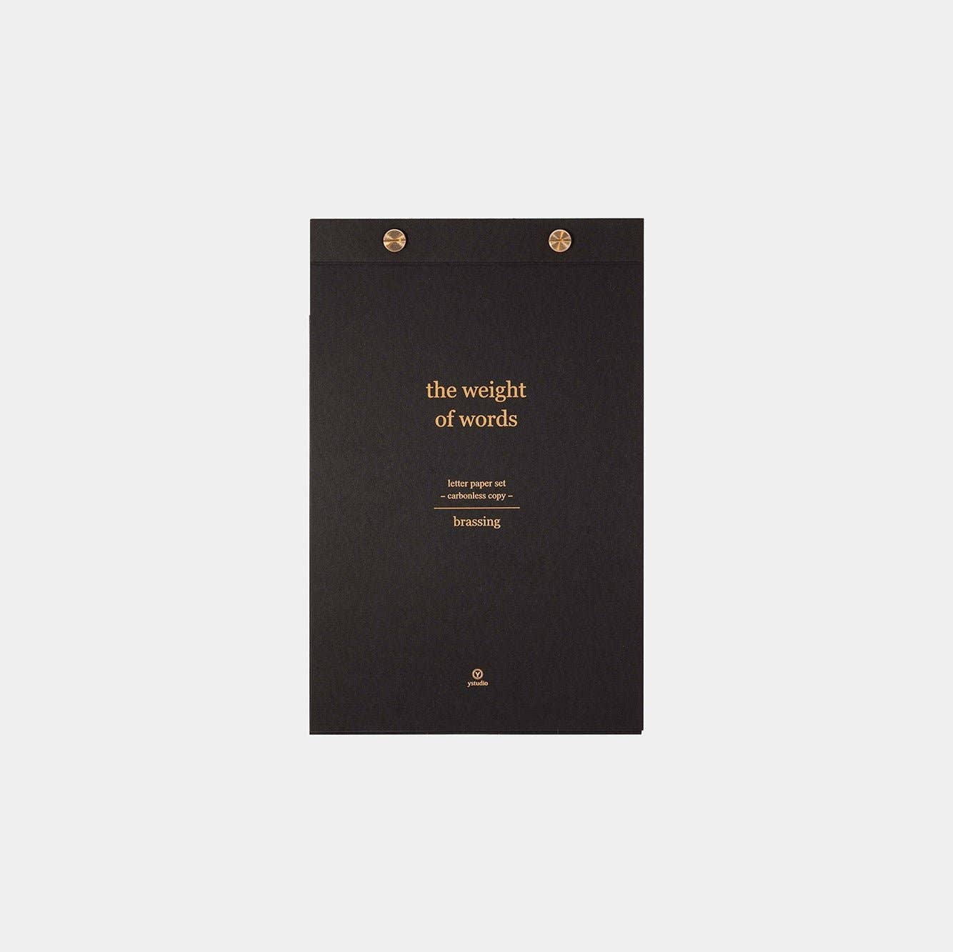 Ein schwarzer ENTREPÔT - YSTUDIO Klassisches Reflect-Briefpapier-Set Notizblock mit goldenem "the weight of words"-Text und zwei goldenen Bindungsklammern steht mittig auf weißem Untergrund - eine raffinierte Ergänzung zu jedem Briefpapier-Set von ENTREPÔT.