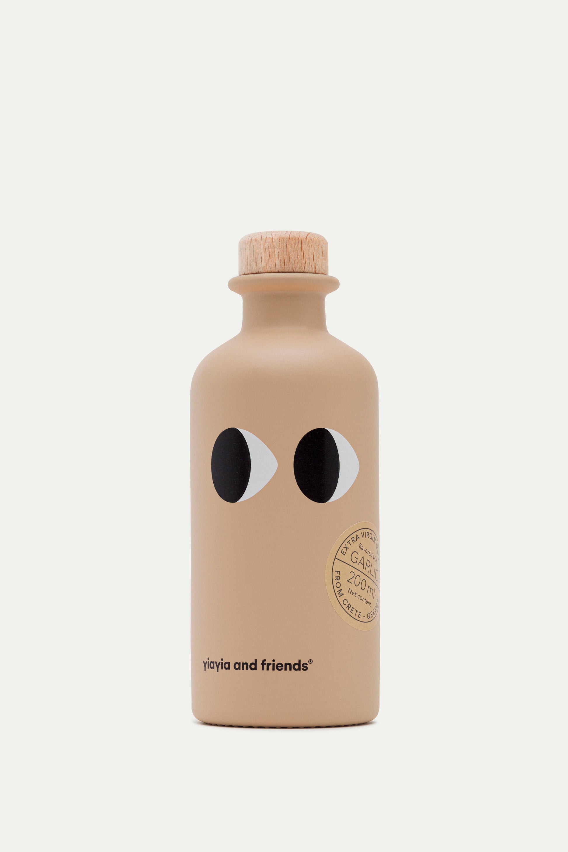 Die beigefarbene Flasche mit Holzdeckel zeigt zwei schwarz-weiße Augen, einen runden Goldstempel und ein Etikett mit der Aufschrift "yiayia and friends". Das einzigartige Design enthält "yiayia and friends - Natives Olivenöl Extra mit Knoblauch". Auf einfarbig weißem Hintergrund.