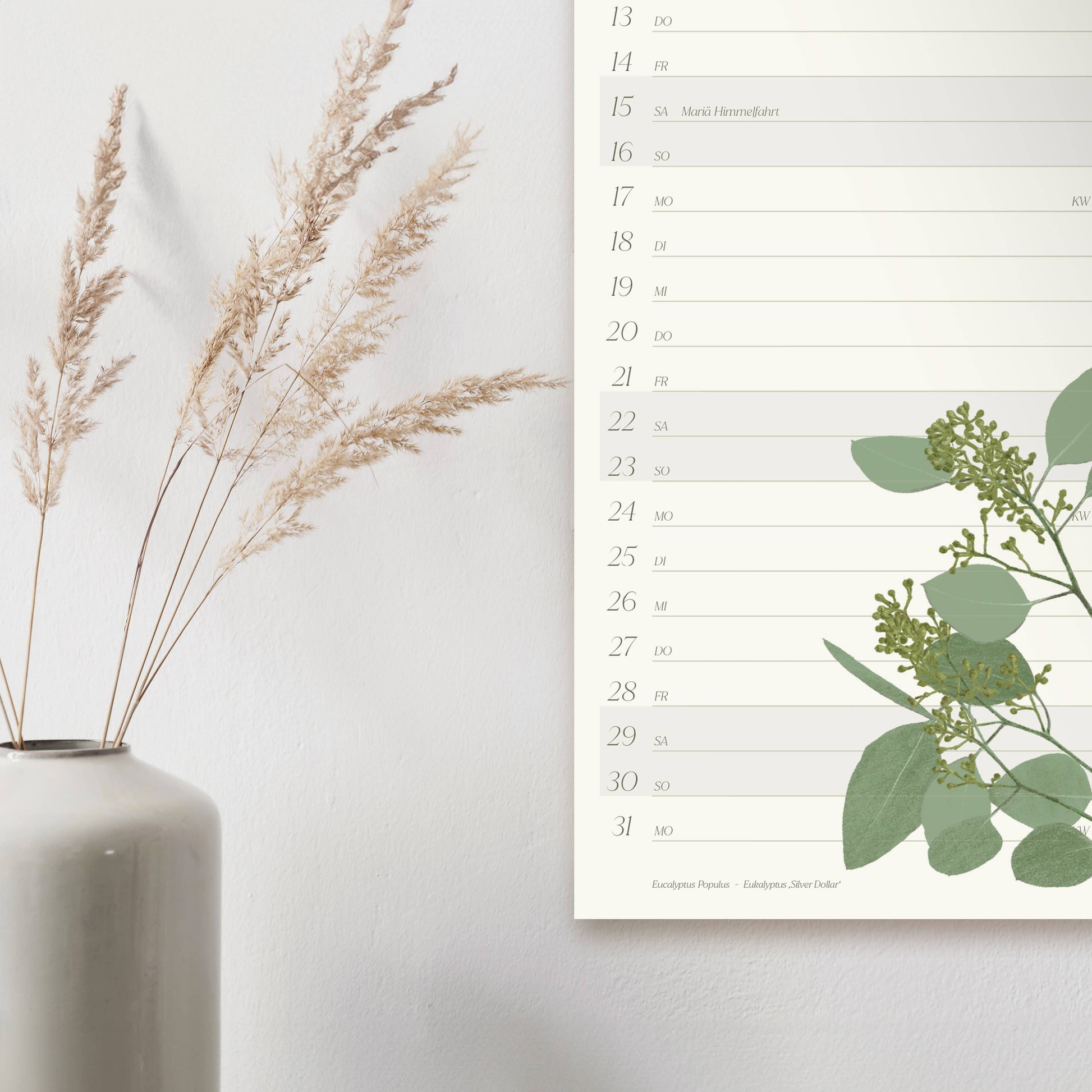 Eine weiße Vase mit getrocknetem Gras steht auf einer weißen Fläche neben dem Wildblumen Atelier - Wandkalender 2026 mit handgezeichneten Blumen, mit Schreibfläche für jedes Datum und gedruckt auf FSC-zertifiziertem Naturpapier.