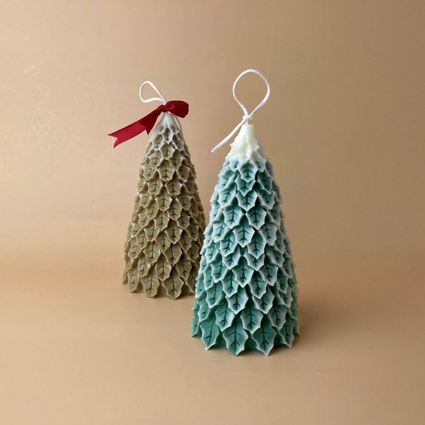 SOULCandle - Weihnachtskerzen von SOULCandle: Zwei handgefertigte Sojawachs-Kerzen im Tannenbaum-Design, grün/weiß und beige mit rotem Band, strukturiert und mit weißen Dochten vor neutralem Hintergrund.