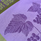 Nahaufnahme des Garnier Thiebaut Küchenhandtuchs Fraisier mit einem lebendigen Weinrebenmuster mit violetten Trauben und großen Blättern auf Lavendel, eingefasst mit grünen und gelben Traubenmotiven – ein elegantes Küchenhandtuch von Garnier Thiebaut.