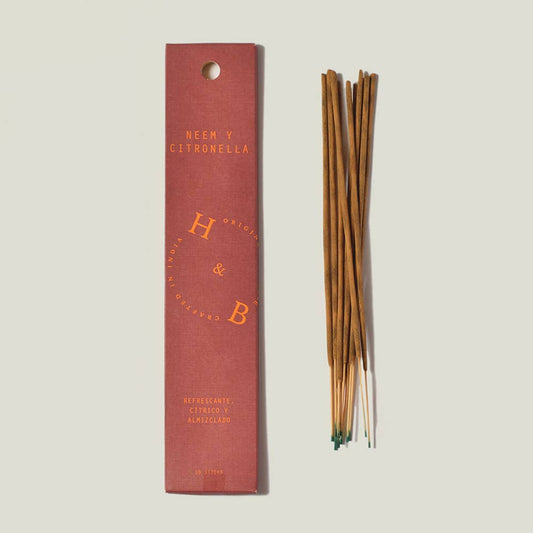 Eine rosafarbene rechteckige H&B-Weihrauchdose mit der Aufschrift "Neem- und Citronella-Weihrauch (gegen Mücken)" liegt neben einem Bündel brauner Räucherstäbchen mit grünen Spitzen, die ordentlich auf einem hellen Hintergrund angeordnet sind.