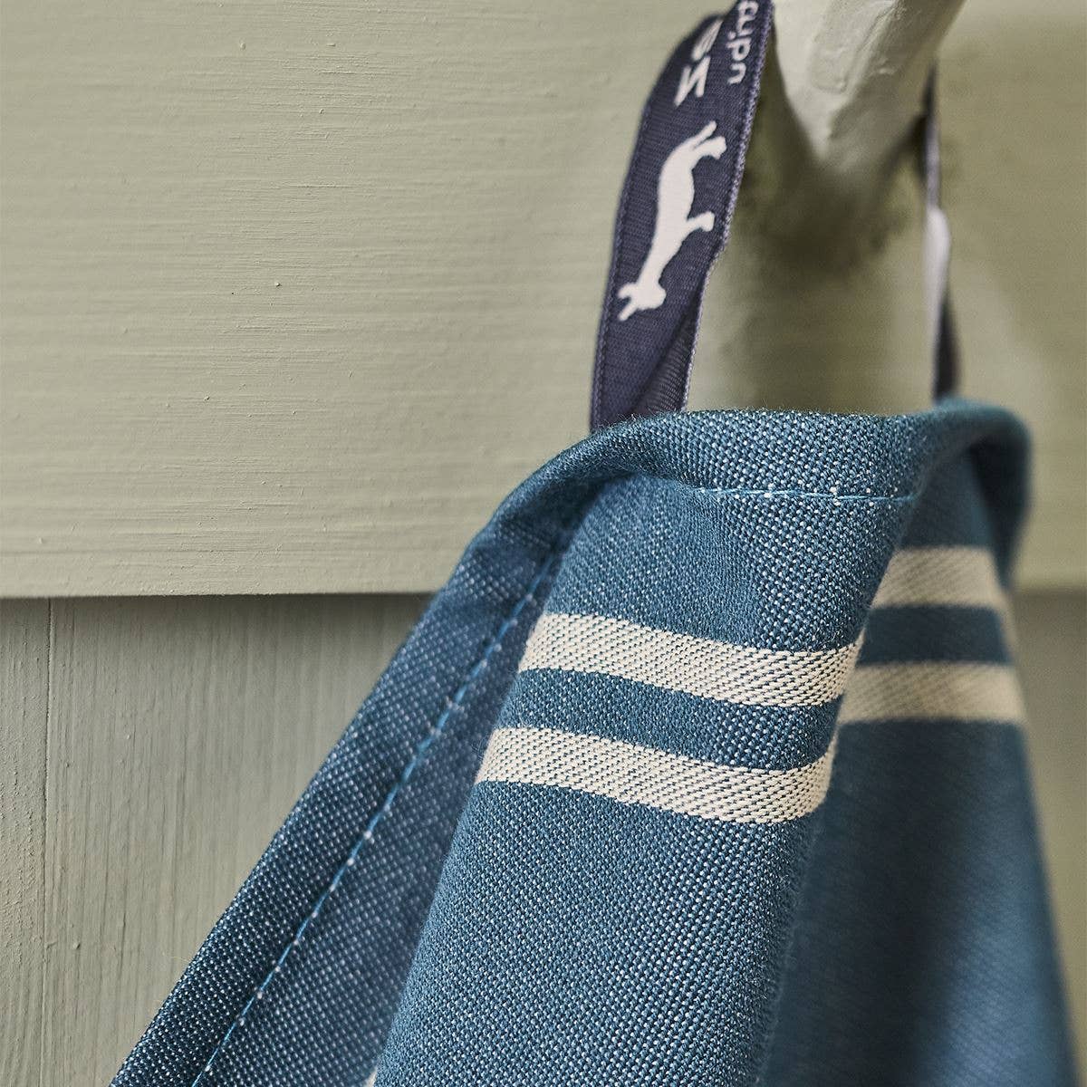 Nahaufnahme des Torchon Jacquard Coeur Vallée Blau von Sylvie Thiriez, ein blaues Geschirrtuch aus 100% Baumwolle mit weißen Streifen, das an einer marineblauen Schlaufe mit weißem Hundemotiv an einem Haken auf hellgrünem Holz hängt.