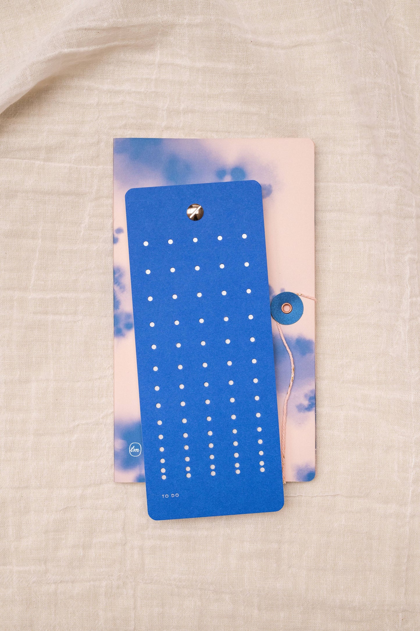 Eine Tinne Mia - Aufgabenliste - True Blue To-Do-Listenkarte von Tinne Mia, nachhaltig produziert aus FSC-Papier, liegt auf einem pastellfarbenen Notizbuch mit Wolkendesign auf strukturiertem beigem Stoff.