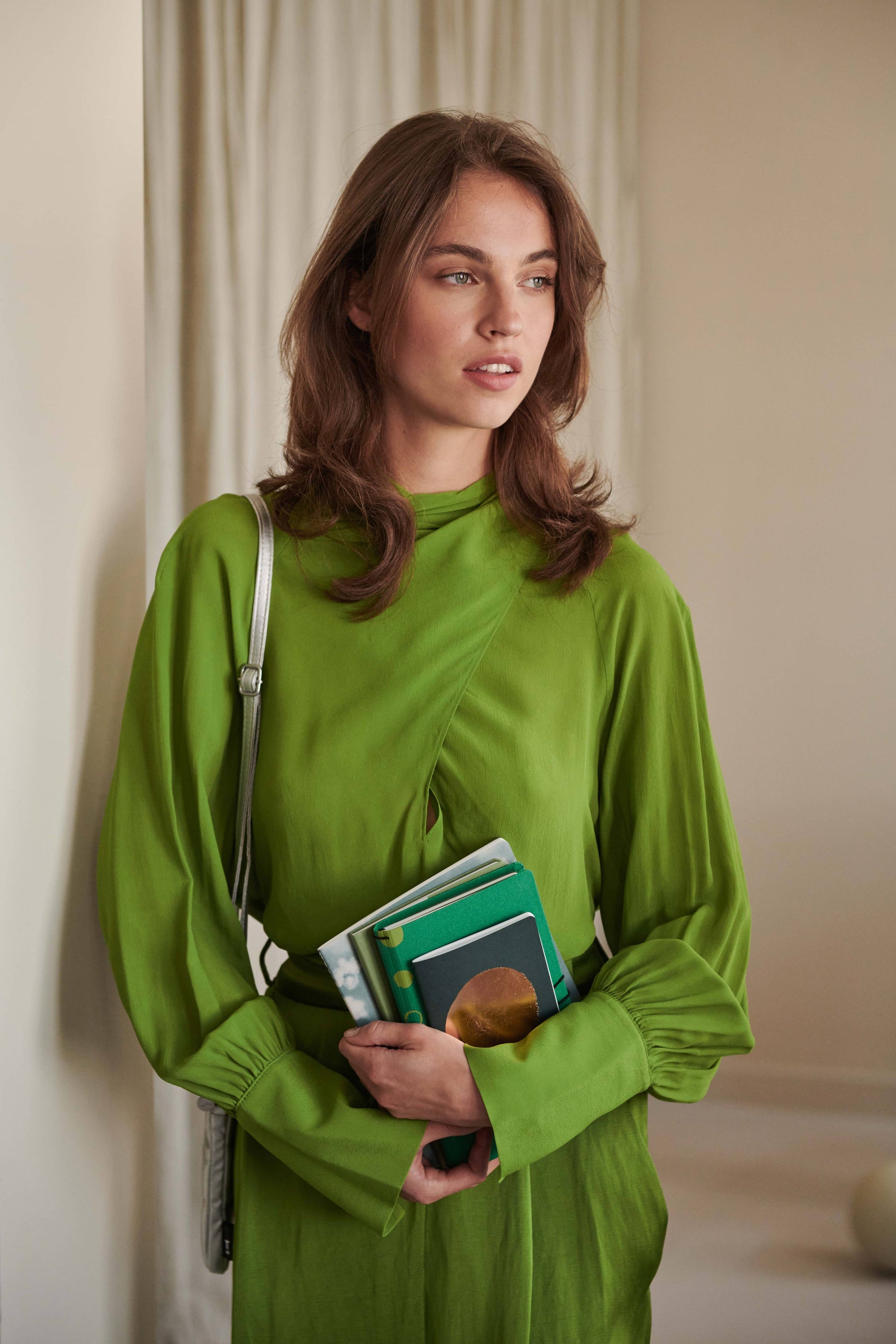 Eine Frau in einem hellgrünen Kleid steht in einem Innenraum und hält mehrere Tinne Mia Notizbücher mit Knopf - Leinen - Smaragd (FSC Mix) in ihren Armen. Sie hat hellbraunes Haar und blickt in einem schwach beleuchteten Raum nachdenklich zur Seite.