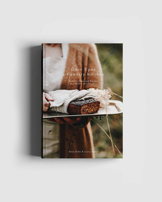 Buchcover für Cozy Publishing – „Es war einmal eine Landhausküche“ von Cozy Publishing. Es zeigt eine Frau mit Schwarzbrot auf einem Leinentuch und weckt so rustikalen Charme. Der unscharfe Hintergrund im Freien verleiht dieser einladenden kulinarischen Geschichte Wärme.