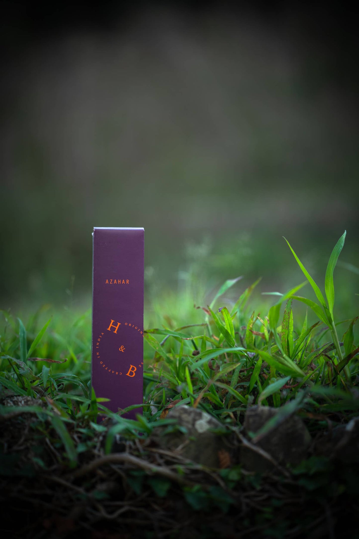 Eine lilafarbene rechteckige Dose mit der Aufschrift H&B Räucherstäbchenhalter Azahar Natürlicher Orangenblüten-Weihrauch steht aufrecht auf grünem Gras vor einem verschwommenen natürlichen Hintergrund, der Noten von Orangenblüte und Sandelholz suggeriert.