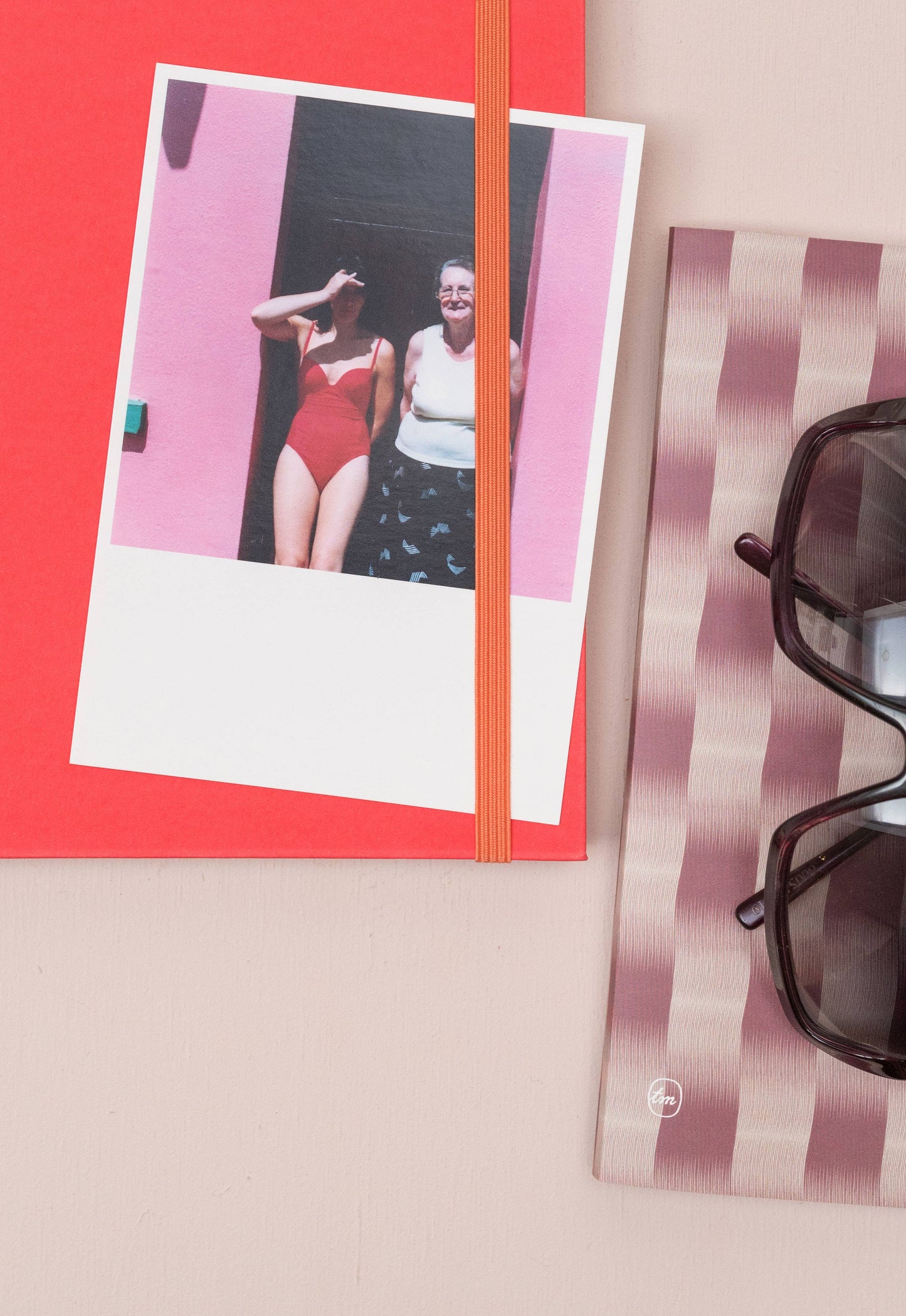 Auf dem Tinne Mia Notizbuch mit japanischem Verschluss in Cinnamon liegt ein Foto von zwei Personen - eine im roten Badeanzug, die andere in weißem Hemd und Shorts. In der Nähe liegt eine Sonnenbrille auf einem rosa gestreiften Tuch über einer hellbeigen Fläche.