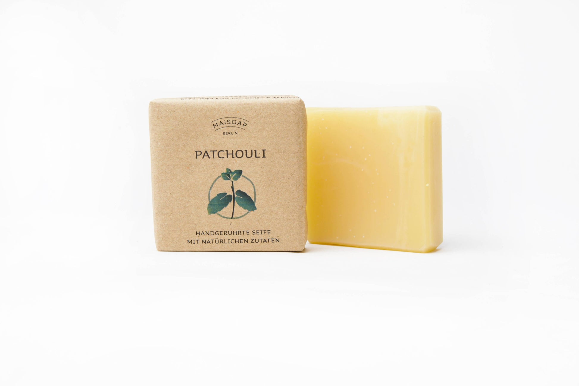 Ein gelber Riegel "Patchouli Seife" von maisoap berlin liegt neben der braunen Papierverpackung mit der Aufschrift "Patchouli & Zitronengras", die eine einfache Patchouli-Blatt-Illustration und einen deutschen Text auf weißem Hintergrund zeigt.