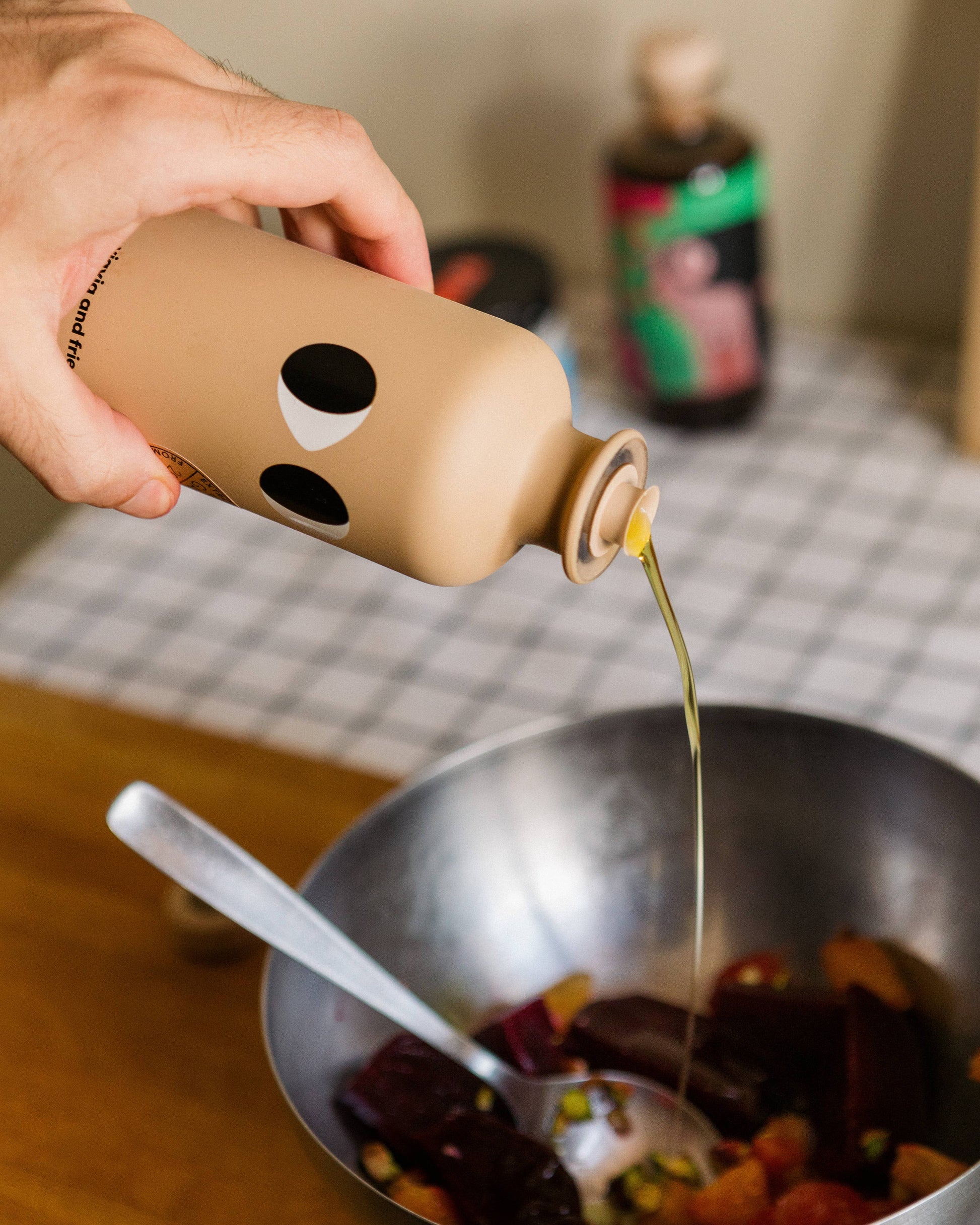 Eine Hand gießt yiayia and friends - Natives Olivenöl Extra mit Knoblauch aus einer hellbraunen Flasche auf einen Gemüsesalat in einer Metallschüssel, mit einem Löffel darin und verschwommenen Flaschen im Hintergrund.