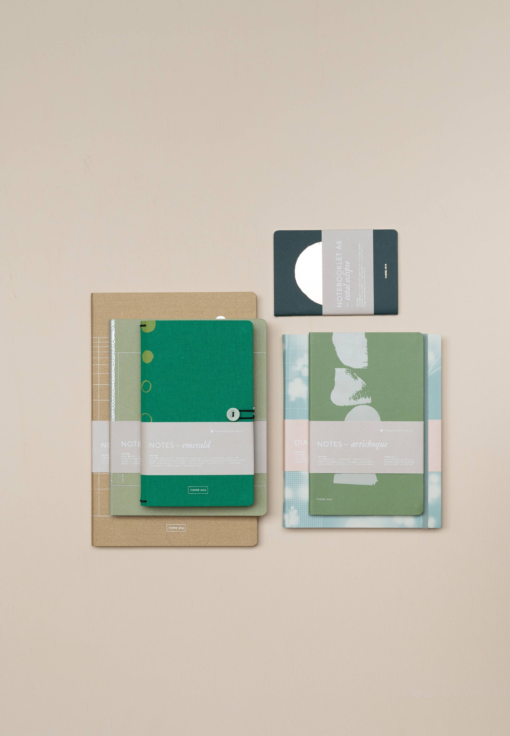 Ein Flat-Lay von vier Tinne Mia Notizbuch mit Knopf in Smaragd, FSC Mix zertifiziert, mit minimalistischen Leinen-Covern in Grün- und Brauntönen auf beigem Hintergrund - ideal als Bullet Journal oder Tagebuch.