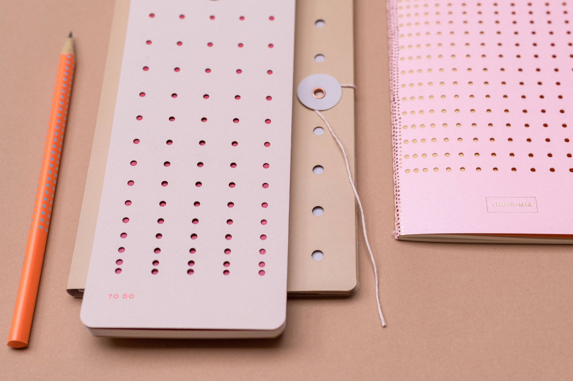 Ein Bleistift und drei Tinne Mia - Aufgabenliste - Mellow Rose Notizblöcke von Tinne Mia, hergestellt aus FSC®-zertifiziertem Papier, in Rosa und Beige mit perforierten Deckeln mit kreisförmigen Löchern, angeordnet auf einer hellbraunen Oberfläche.