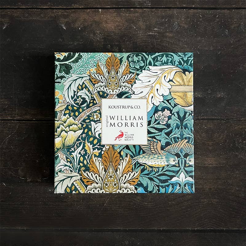 Eine quadratische Koustrupco-Box mit botanischen und Vogelmotiven in Grün-, Gelb- und Cremetönen sitzt auf dunklem Holz und erinnert an die Eleganz des Koustrupco Seidenschal Morris Pimpernel Blau 50 x 50 cm.