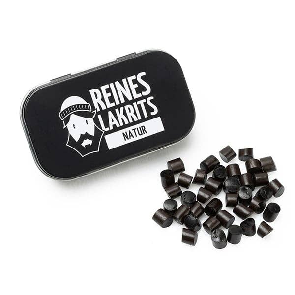 Eine schwarze Dose mit der Aufschrift „LASSE LAKRITS – natürlich lecker – Dose Reines Lakritz Natur“ mit der Grafik eines bärtigen Gesichts wird neben mehreren kleinen, runden Lakritzstückchen aus natürlichen Zutaten auf einer weißen Fläche platziert.