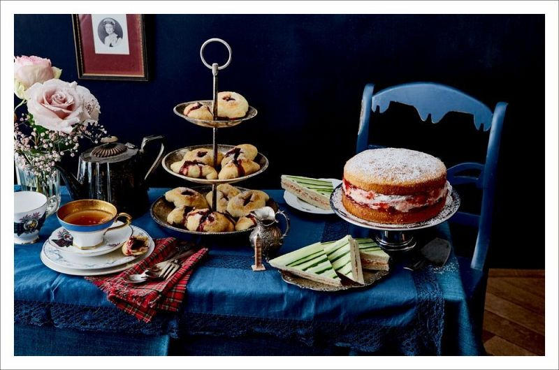 Das Tischset "London Lovestory" vom Hölker Verlag mit blauem Tuch, Stufentablett mit Scones, Biskuit, Gurkensandwiches, Teekanne, Teetasse, Blumen in einer Vase und blauem Stuhl bringt Londoner Romantik in die Teestunde.