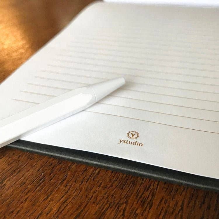 Ein weißer Stift liegt auf einem aufgeschlagenen, linierten ENTREPÔT ystudio Notizbuch mit dem Schriftzug "ystudio" am unteren Rand der Seite. Das ENTREPÔT - YSTUDIO - Klassisches Reflect-Briefpapier-Set liegt auf einer Holzunterlage.