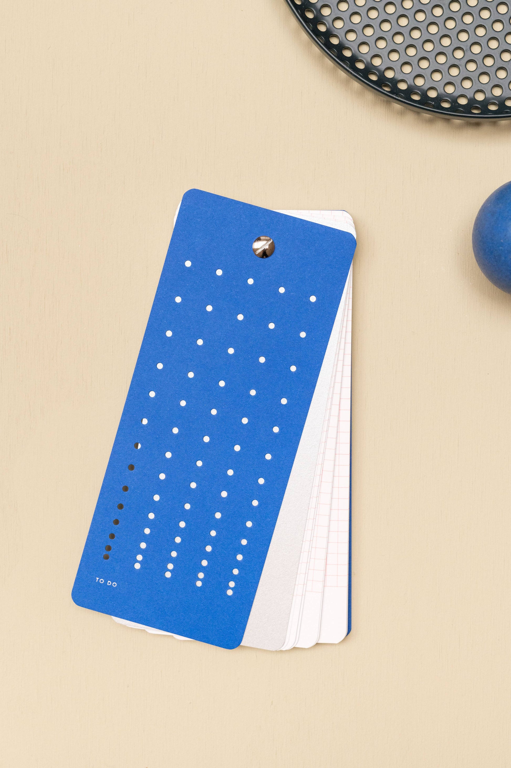 Die Tinne Mia - Aufgabenliste - True Blue, ein nachhaltig hergestellter Notizblock aus FSC-Papier mit To Do-Liste und weißem Punktraster, liegt offen auf einer beigen Fläche neben einem schwarzen perforierten Tablett und einem blauen runden Objekt.