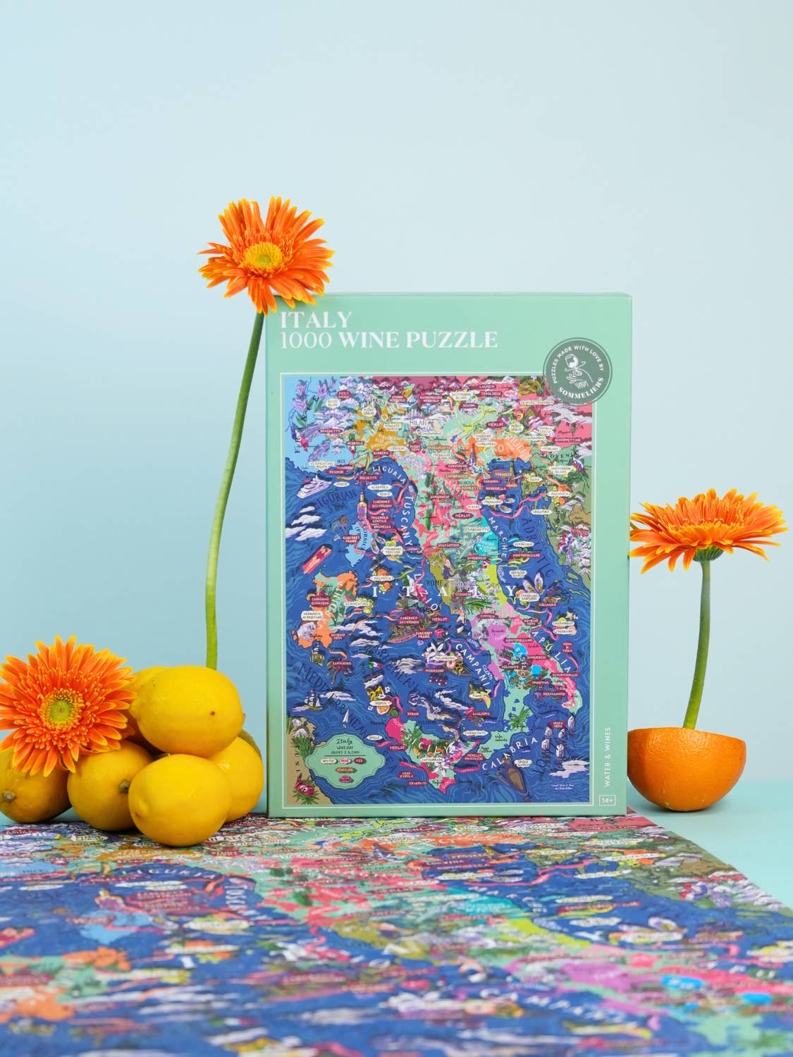 Das Water & Wines - Wein Puzzle - Italien von Water & Wines, ein lebendiges 1000-teiliges Puzzle der italienischen Weinregionen, wird zwischen orangefarbenen Blumen und Zitronen gezeigt, wobei ein Teil des fertigen Puzzles vor einem hellblauen Hintergrund zu sehen ist.