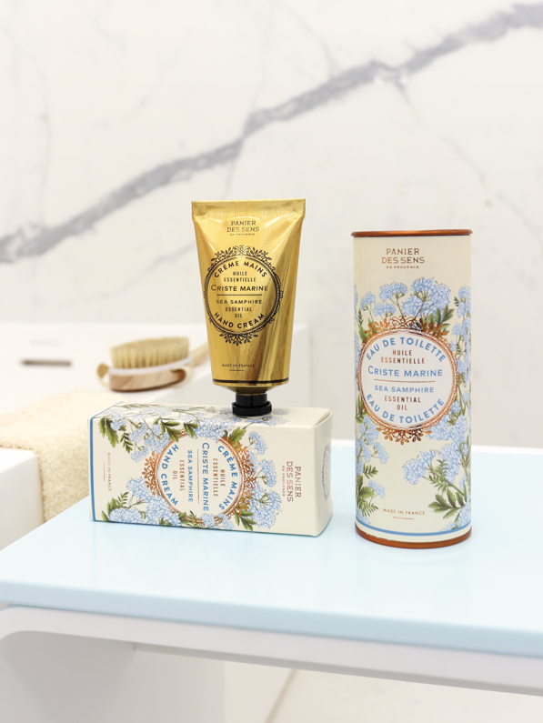 Ein Riegel Panier Des Sens Pflanzliche Seife | Meerfenchel 150 g liegt auf einer blauen Fläche neben der Schachtel, im Hintergrund eine Bürste mit weichen Borsten und ein weißes Handtuch.