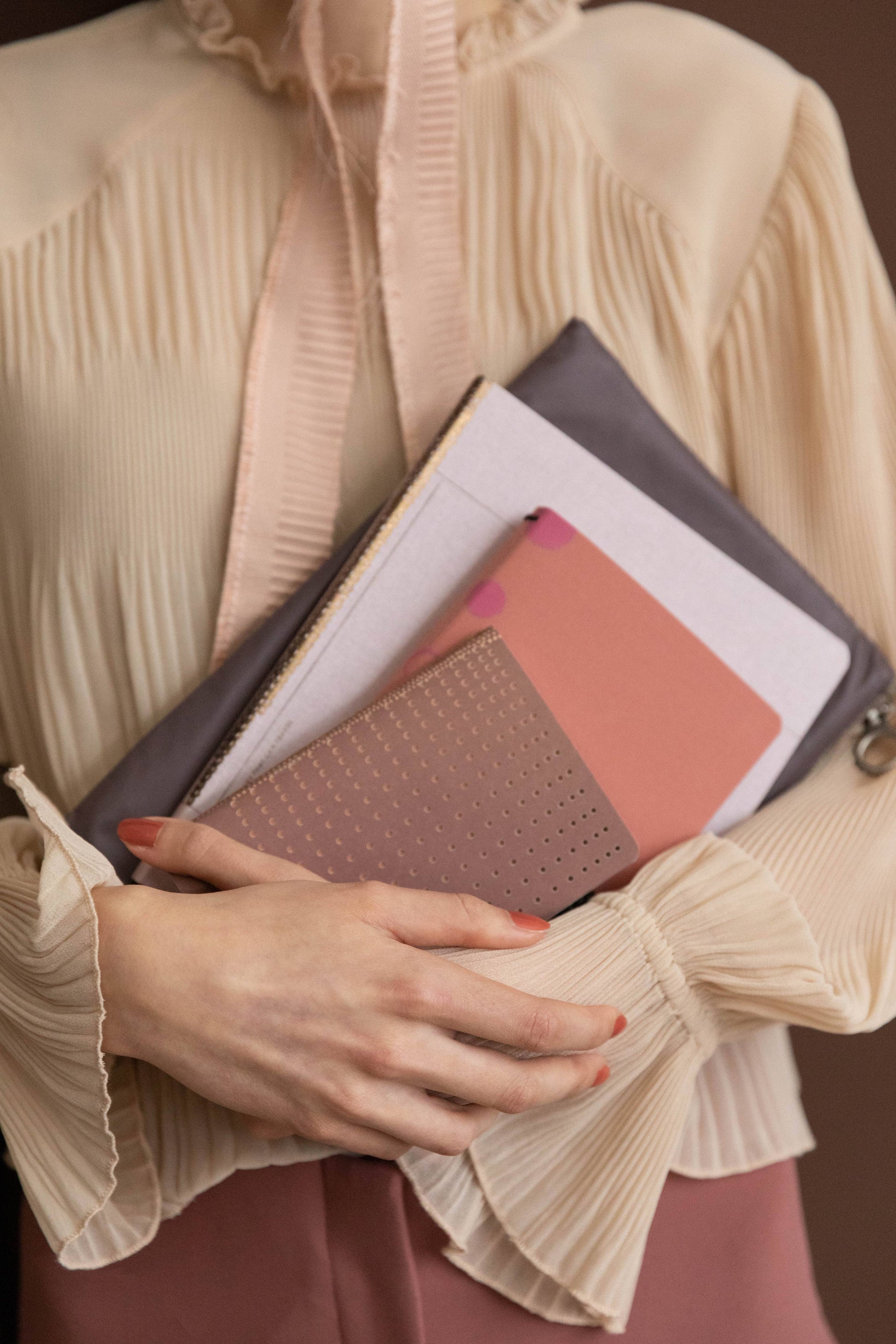 Eine Person in cremefarbener Plisseebluse hält Tinne Mias Notizbuch mit Knopf - Leinen - Rose Dawn FSC Mix zusammen mit passenden rosafarbenen und lilafarbenen Mappen in der Hand, ihre ordentlich manikürten Hände sind sichtbar.