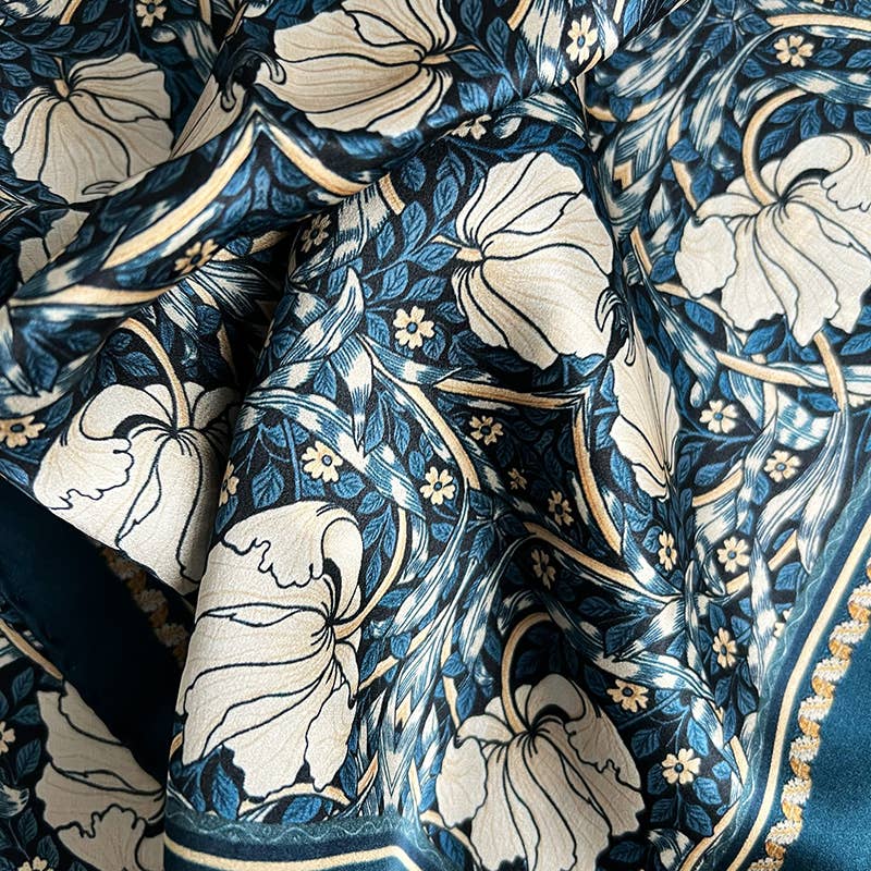 Eine Nahaufnahme des von William Morris inspirierten Koustrupco | Seidenschal Morris Pimpernel Blau 50 x 50 cm zeigt große cremefarbene Blumen, dunkelgrüne Blätter und goldene Details auf einem dunkelblauen Hintergrund; der Seidenschal wirkt weich und leicht gefaltet.