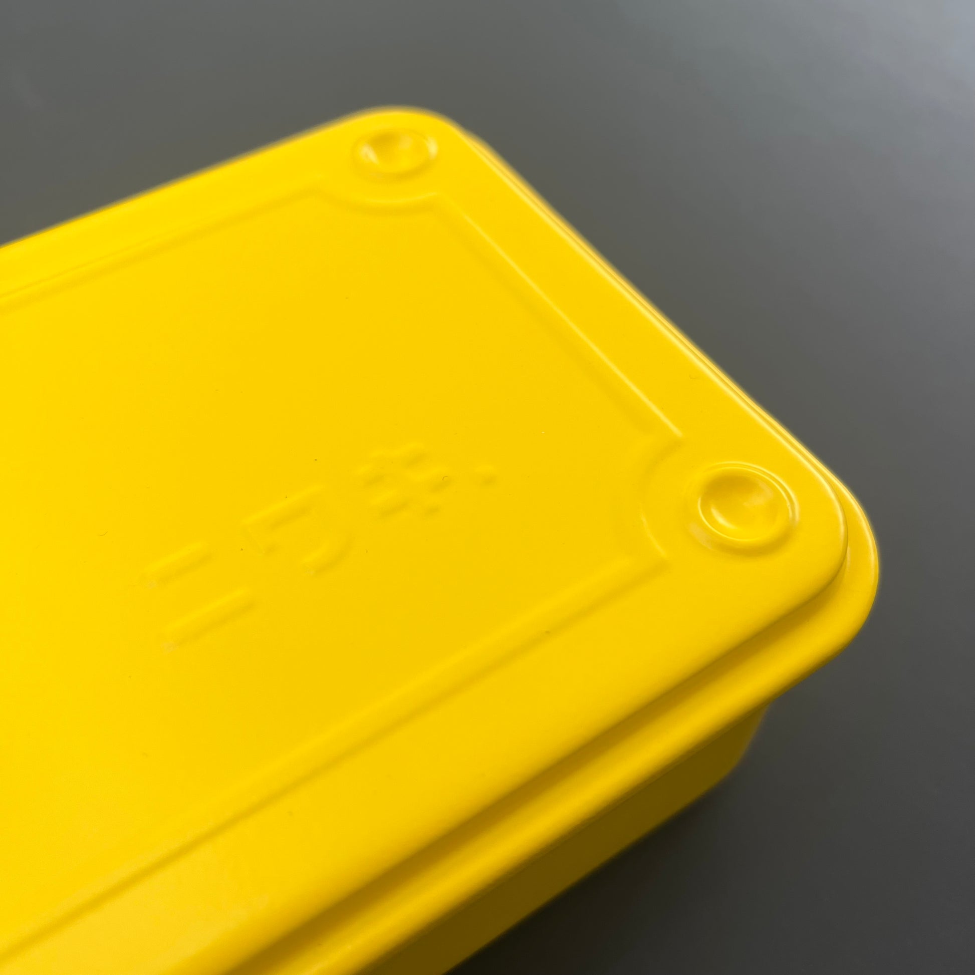Eine Nahaufnahme der leuchtend gelben Niwaki T-Type Toolbox von Niwaki mit abgerundeten Ecken, einem geprägten rechteckigen Deckelrand und eingekerbten japanischen Schriftzeichen – ideal für Gärtner und Heimwerker – vor einem grauen Hintergrund.