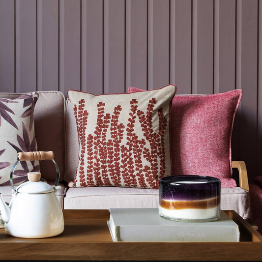 Ein gemütliches Wohnzimmer mit einem rosa Sofa, dem bestickten Kissen mit Kräutermotiv Persimone von Riva Home, einer weißen Teekanne mit Holzgriff, einer Glaskerze und gestapelten Büchern auf einem Holztablett vor einer malvenfarbenen getäfelten Wand.