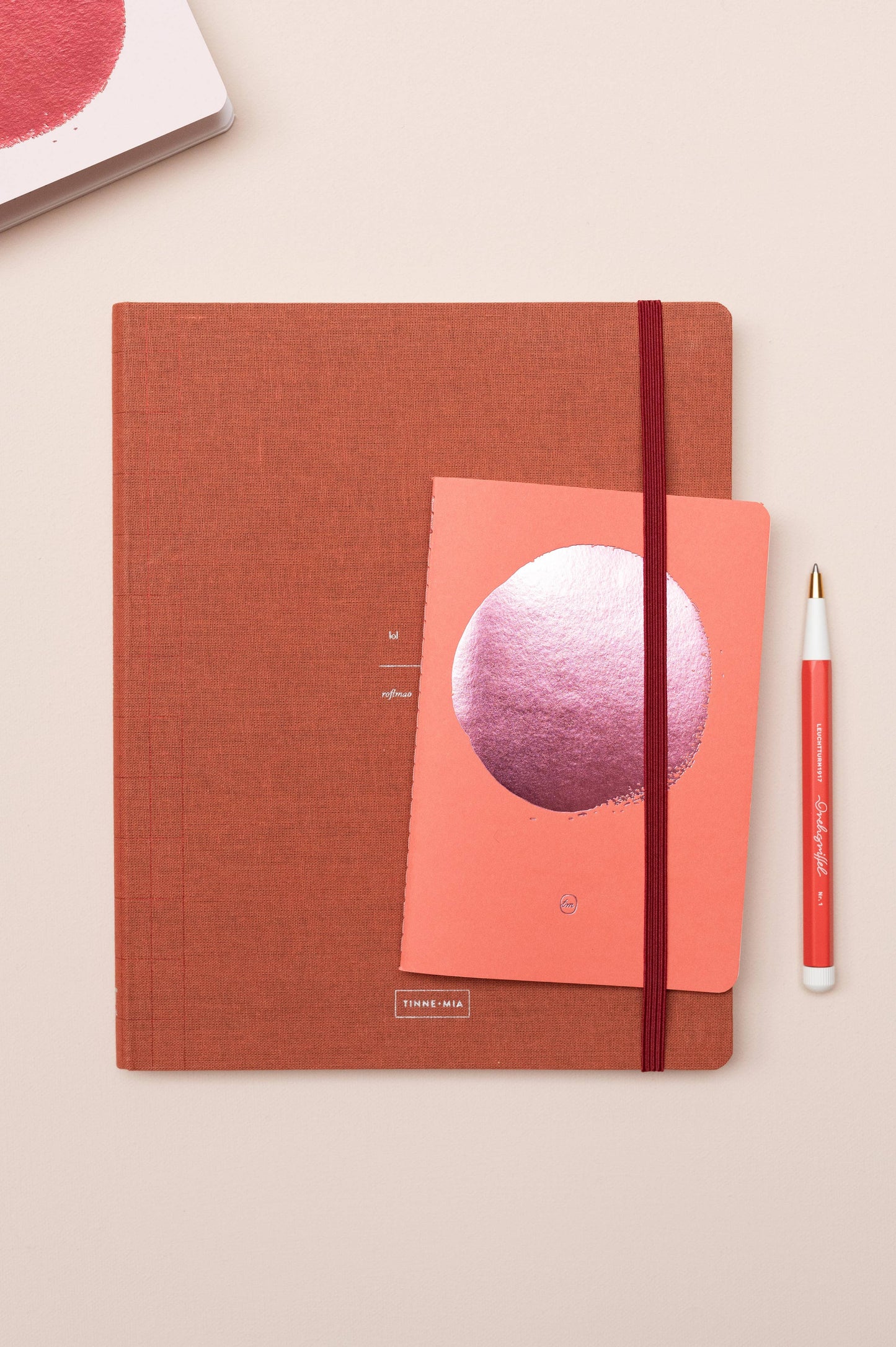 Ein Flat-Lay mit dem Tinne Mia Bullet Journal A6 in Flamingo (FSC Mix), einem kleineren korallenroten Notizbuch mit metallic-pinkem Kreisdesign und einem weiß-korallenroten Stift auf beiger Oberfläche - ideal für Notizen auf FSC-zertifiziertem Papier.