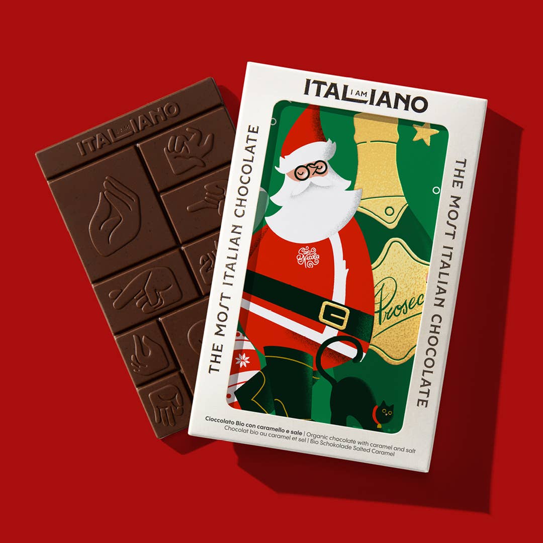 Ein Riegel I AM ITALIANO - Prosecco Bio-Schokolade mit Karamell und Salz mit Handzeichen sitzt auf einer festlichen Verpackung mit einem Weihnachtsmann, der eine Prosecco-Flasche hält, und "ITALIANO, THE MOST ITALIAN CHOCOLATE" auf rotem Hintergrund.