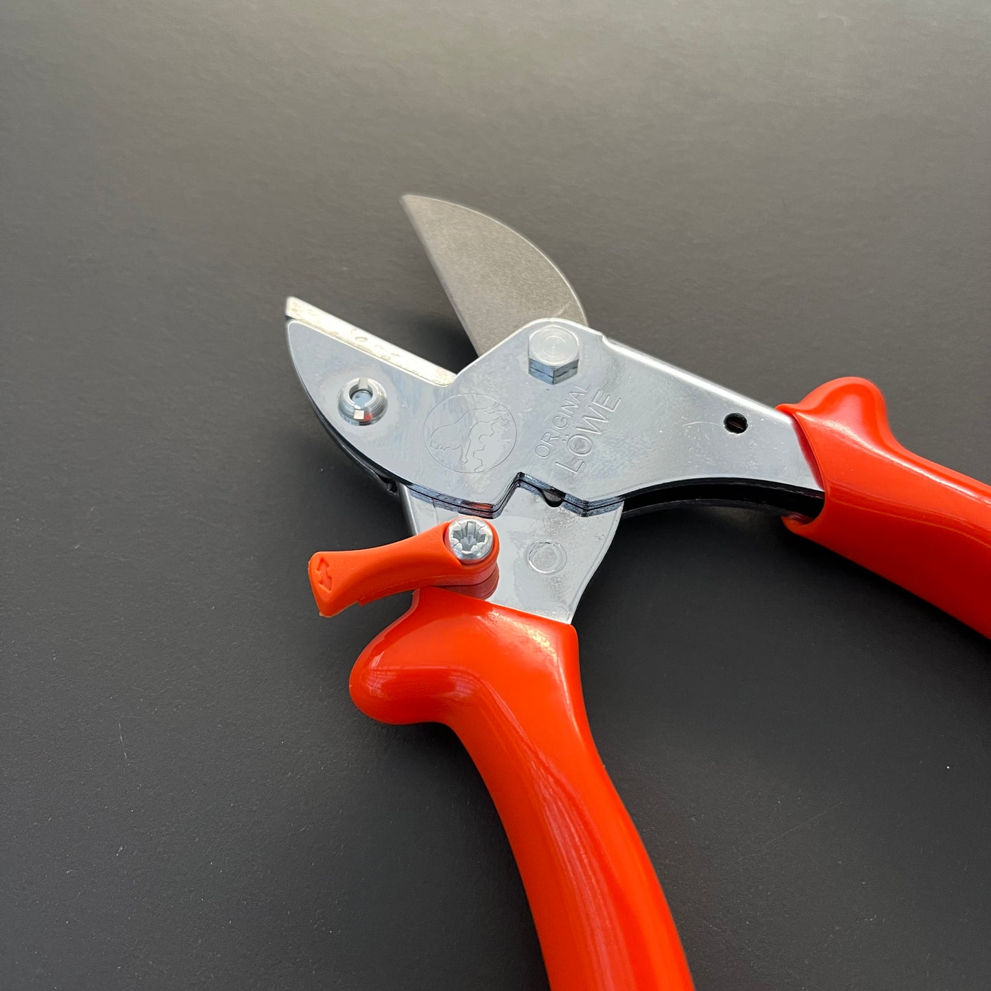 Auf einer dunklen, glatten Fläche ist eine Löwe Original Löwe 5 - Classic Easy Cut Gartenschere mit leuchtend orangefarbenen Griffen und einer silberglänzenden Klinge mit Amboss-Technik abgebildet.