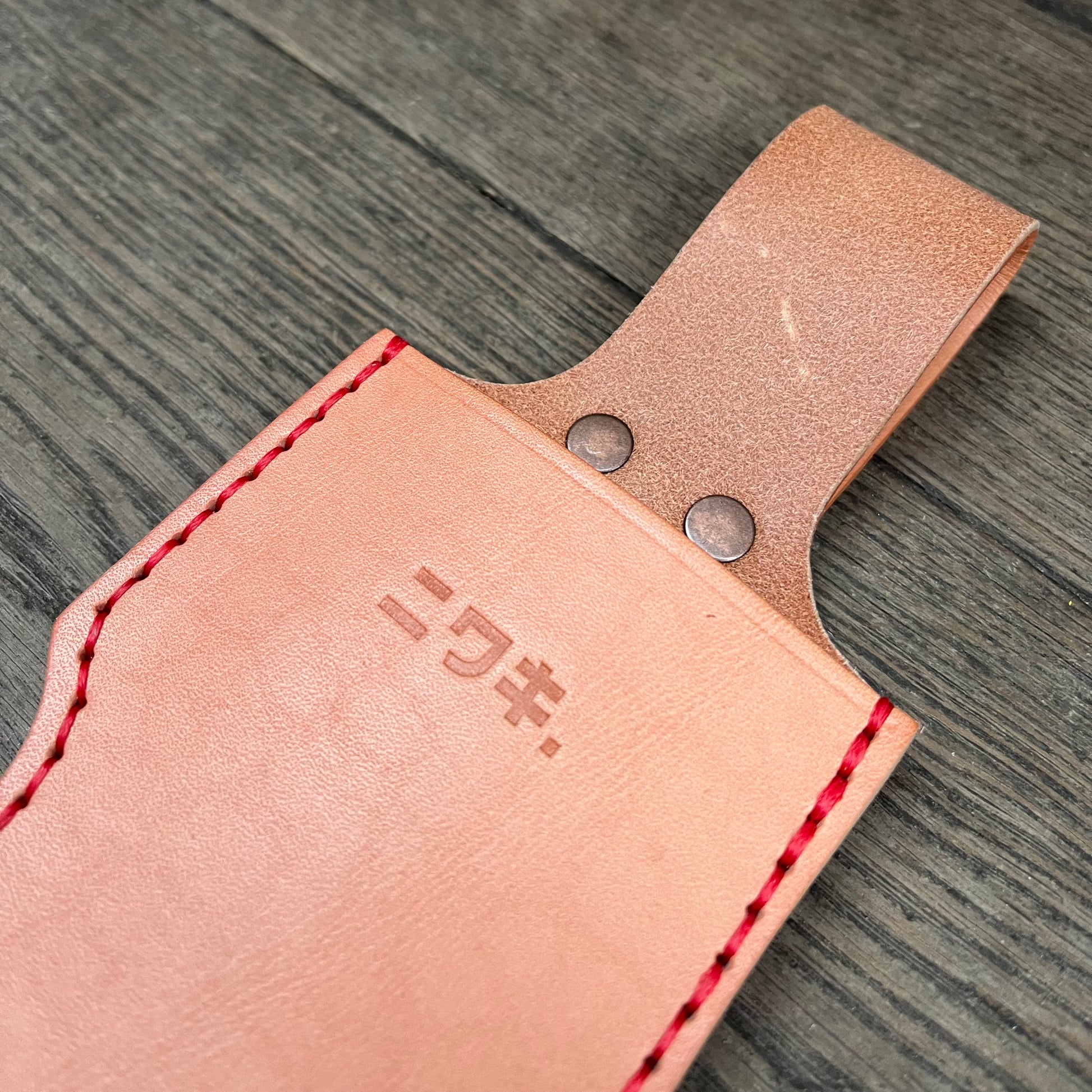 Eine Nahaufnahme des Niwaki Single Holsters, einer hellbraunen Lederscheide von Niwaki mit roten Nähten und zwei Metallnieten. Japanische Schriftzeichen sind in das Leder eingeprägt, während es auf einer Holzoberfläche liegt.