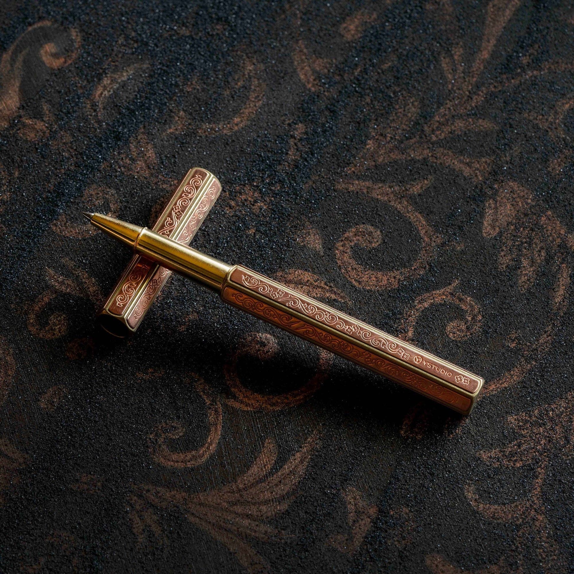 Der ENTREPÔT YSTUDIO Klassische Renaissance KAZARI KANAGU ORCHID Tintenroller aus Gold und Bronze mit aufwendigen Gravuren ruht auf seiner passenden Kappe auf einer dunklen, kunstvoll gemusterten Oberfläche.