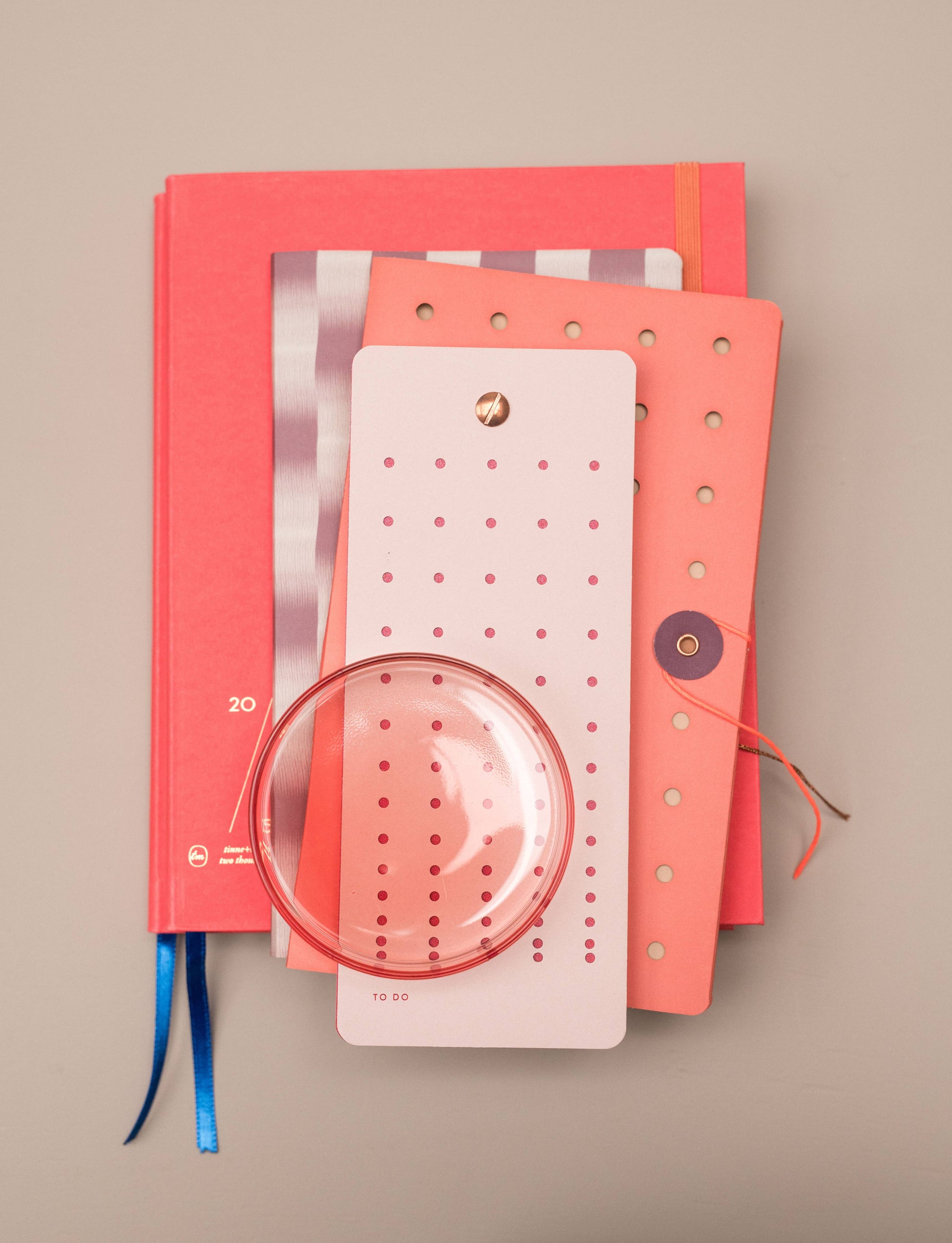 Ein Tinne Mia - Notizbuch mit japanischem Verschluss in Cinnamon von Tinne Mia, fein säuberlich angeordnet auf einer hellbeigen Fläche mit einer runden Lupe und einem rosa gepunkteten Lesezeichen.