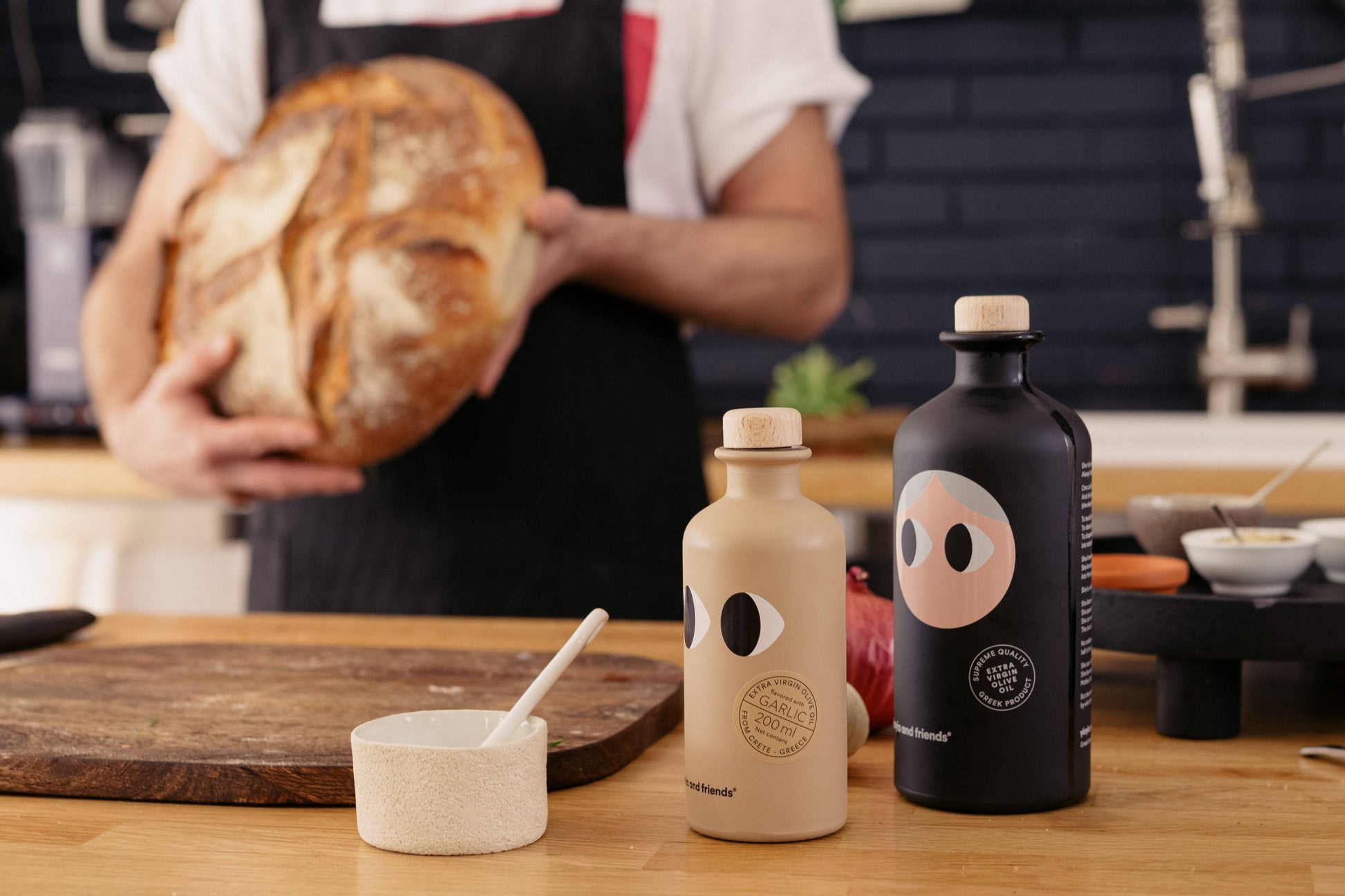 Eine Person in einer Schürze hält rustikales Brot in einer Küche. Im Vordergrund stehen zwei stilvolle Flaschen - eine ist yiayia and friends - Natives Olivenöl Extra mit Knoblauch - und eine kleine Schale mit Löffel auf dem Holztisch.