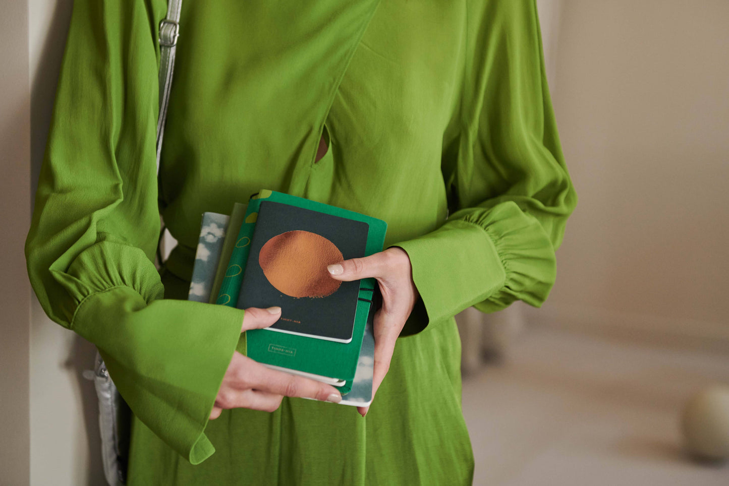 Eine Person in einem leuchtend grünen Kleid hält mehrere bunte Notizbücher, darunter das Tinne Mia Notizbuch mit Knopf aus Leinen in Smaragd (FSC Mix), dicht vor der Brust. Ihr Kopf befindet sich außerhalb des Bildausschnitts.