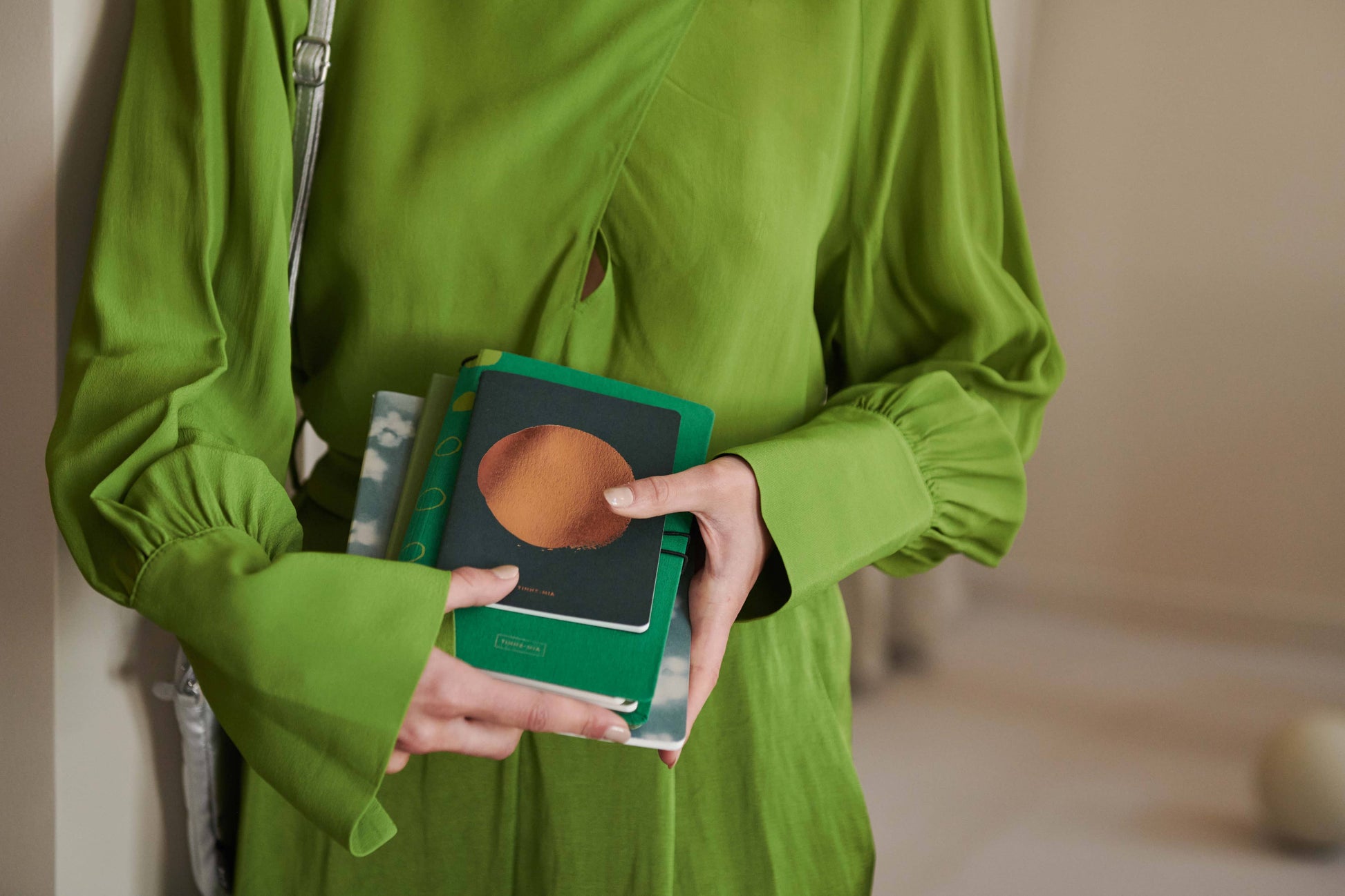 Eine Person in einem leuchtend grünen Kleid hält mehrere bunte Notizbücher, darunter das Tinne Mia Notizbuch mit Knopf aus Leinen in Smaragd (FSC Mix), dicht vor der Brust. Ihr Kopf befindet sich außerhalb des Bildausschnitts.