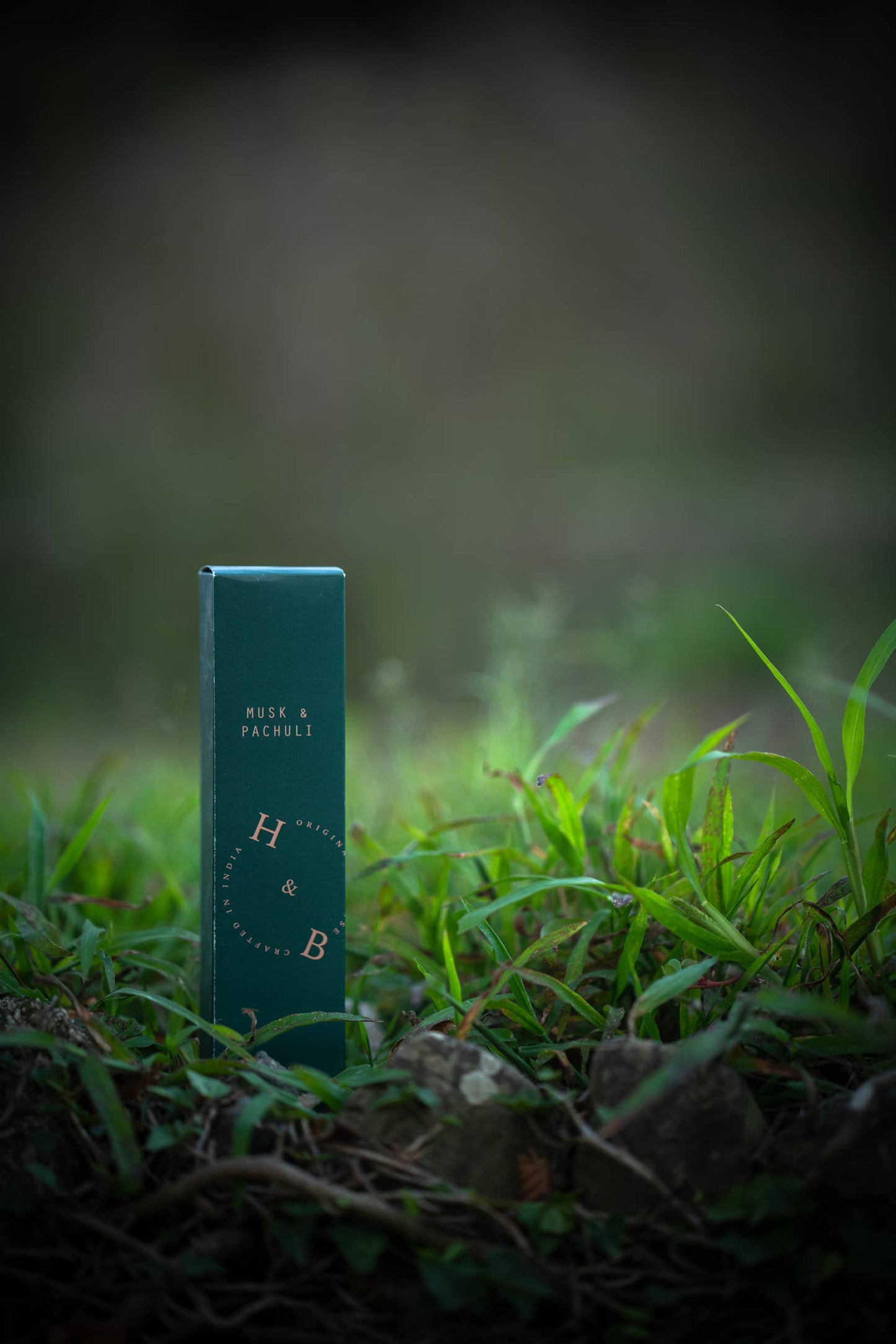Eine grüne, rechteckige Schachtel mit der Aufschrift "H&B Incense - Natürlicher Moschus- und Patchouli-Weihrauch" steht aufrecht zwischen Gras und Steinen, mit einem erdigen Hintergrund, der die reichhaltigen Düfte des natürlichen Moschus- und Patchouli-Weihrauchs von H&B Incense hervorruft.
