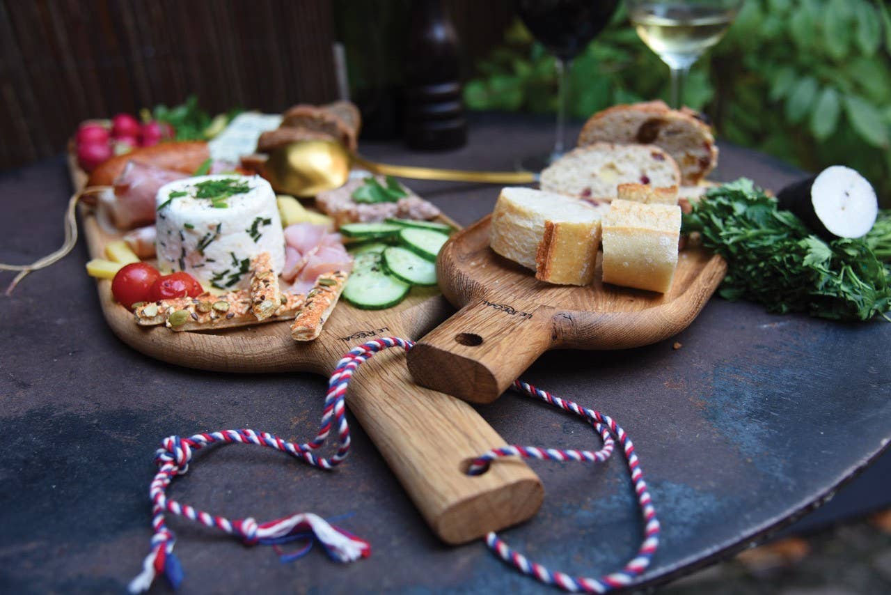 Zwei LE RÉGAL Große Schaufel Aperitifbretter aus französischem Eichenholz mit Käse, Brot, Wurst und Obst stehen auf einem runden Tisch; zwei Weingläser und eine Kordel unterstreichen die nachhaltige Produktion.