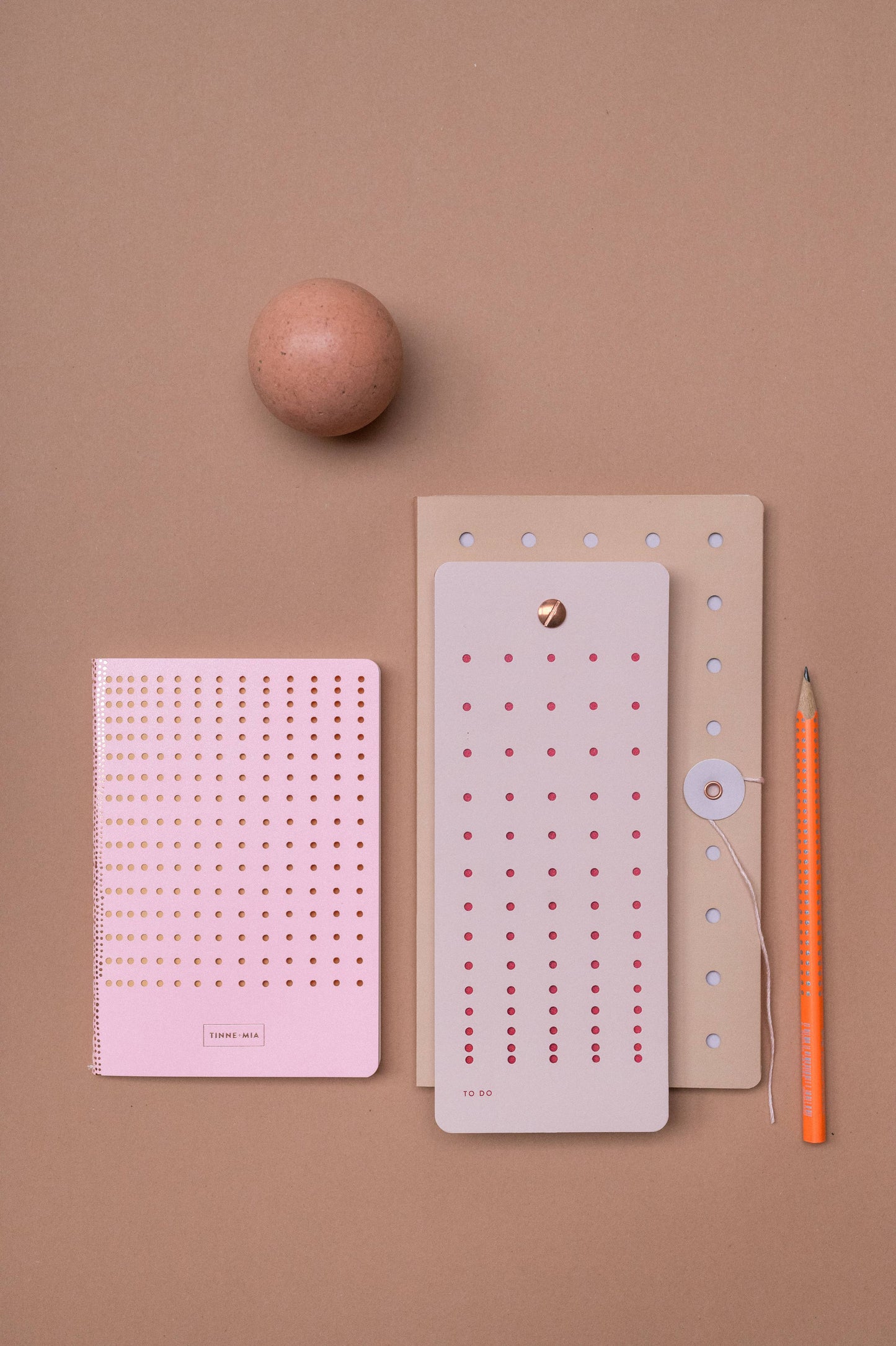 Ein braunes Ei, ein rosafarbener Tinne Mia - Aufgabenliste - Mellow Rose Notizblock von Tinne Mia, ein hellbrauner Ordner, ein großer Listenbuch-Notizblock, ein orangefarbener Bleistift und ein runder Papierverschluss befinden sich auf einem beigen Hintergrund. Alle Papierartikel haben Punkte und sind aus FSC®-zertifiziertem Papier.