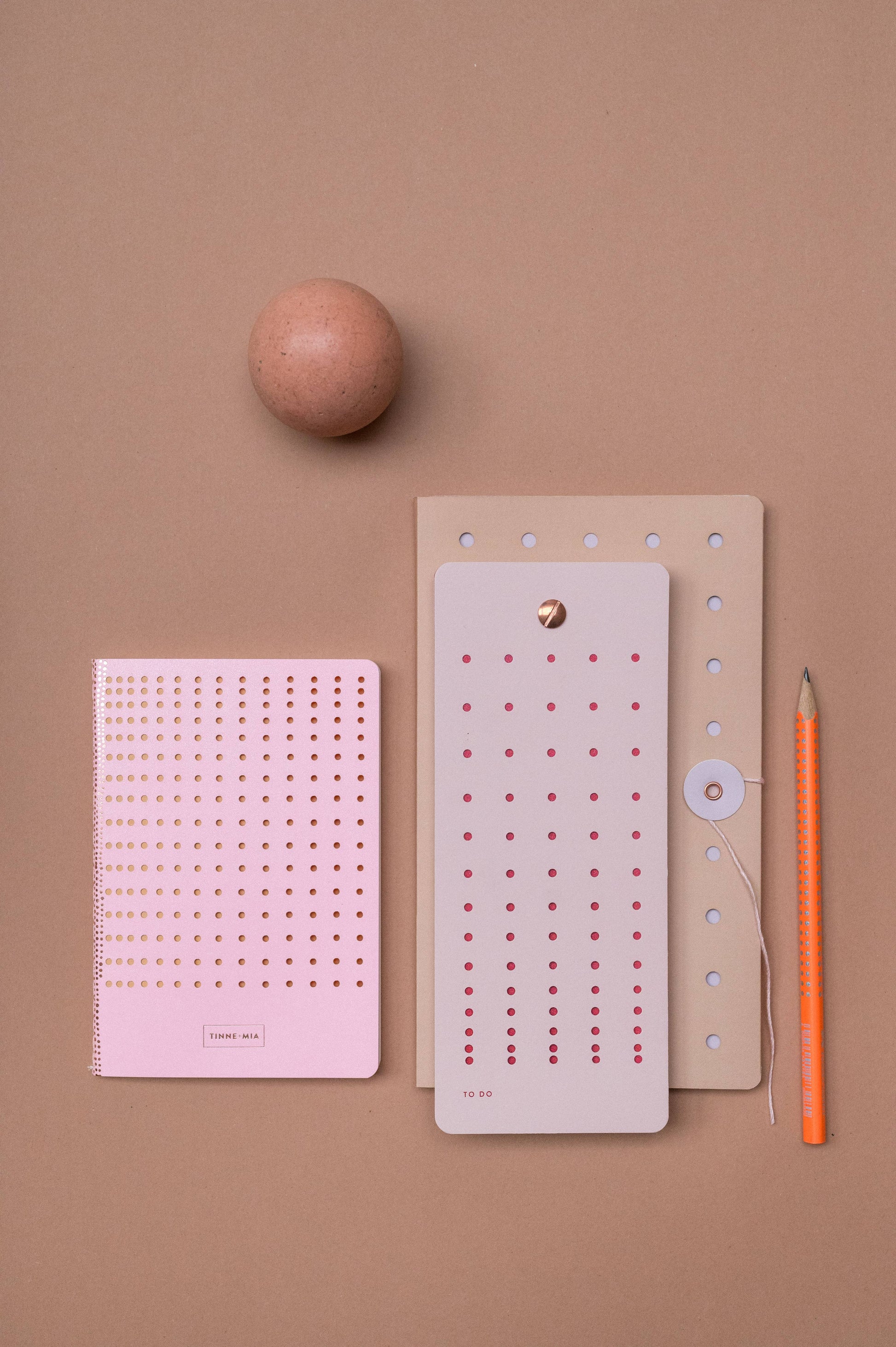 Ein braunes Ei, ein rosafarbener Tinne Mia - Aufgabenliste - Mellow Rose Notizblock von Tinne Mia, ein hellbrauner Ordner, ein großer Listenbuch-Notizblock, ein orangefarbener Bleistift und ein runder Papierverschluss befinden sich auf einem beigen Hintergrund. Alle Papierartikel haben Punkte und sind aus FSC®-zertifiziertem Papier.