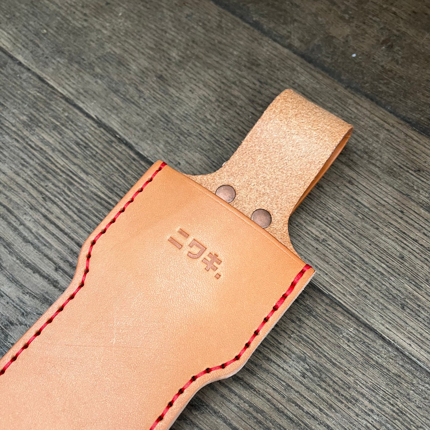 Das Niwaki Long Holster von Niwaki ist eine hellbraune Lederscheide mit roten Nähten und geprägten japanischen Schriftzeichen, ideal für große Scheren. Es verfügt über eine mit Metallnieten gesicherte obere Schlaufe und ruht auf einer Holzoberfläche.