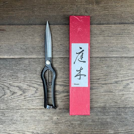 Eine schwarze Niwaki Sentei-Baumschere liegt neben einer roten rechteckigen Niwaki-Schachtel mit japanischer Kalligrafie auf dem Etikett auf einer Holzfläche.