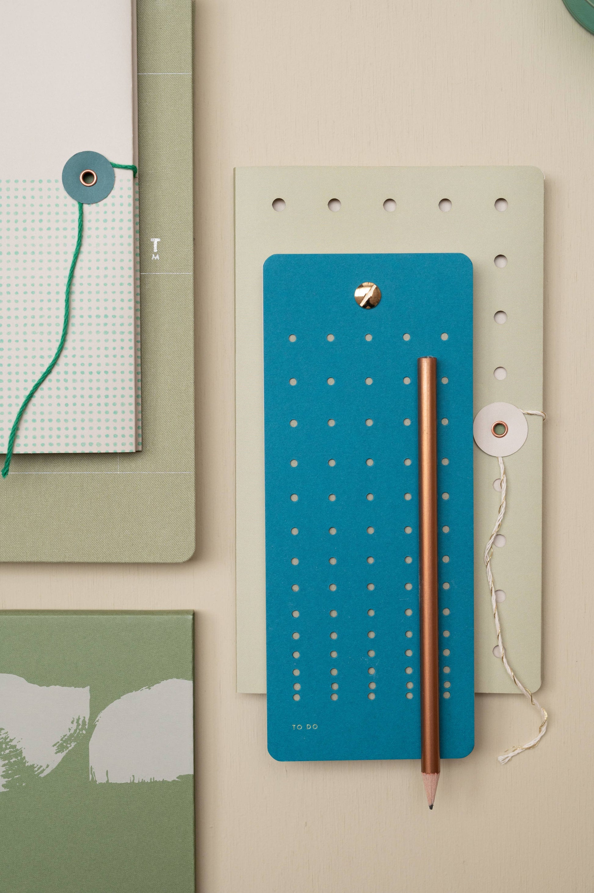 Ein flaches Briefpapier mit Tinne Mia - Aufgabenliste - Blaugrün (FSC®-zertifiziert), einem goldenen Bleistift, grün-blauen Notizbüchern und dekorativen Kordelverschlüssen auf beigem Hintergrund.