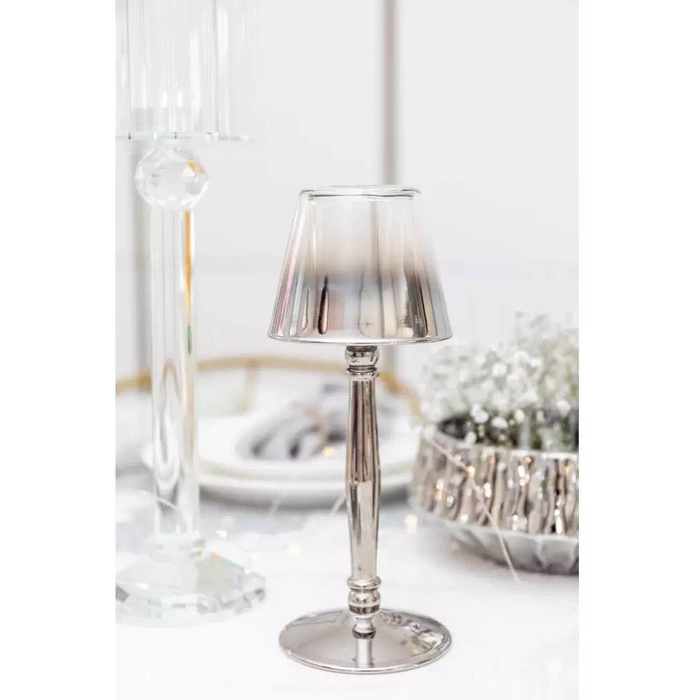 Eine kleine silberne Lampe mit Metallschirm steht auf einer weißen Tischplatte, umgeben von eleganten Glaswaren und dem AULICA - Silberner Kerzenhalter auf Fuß 9,5x9,5x22,5cm von AULICA, neben einer dekorativen Schale und zarten weißen Blumen.