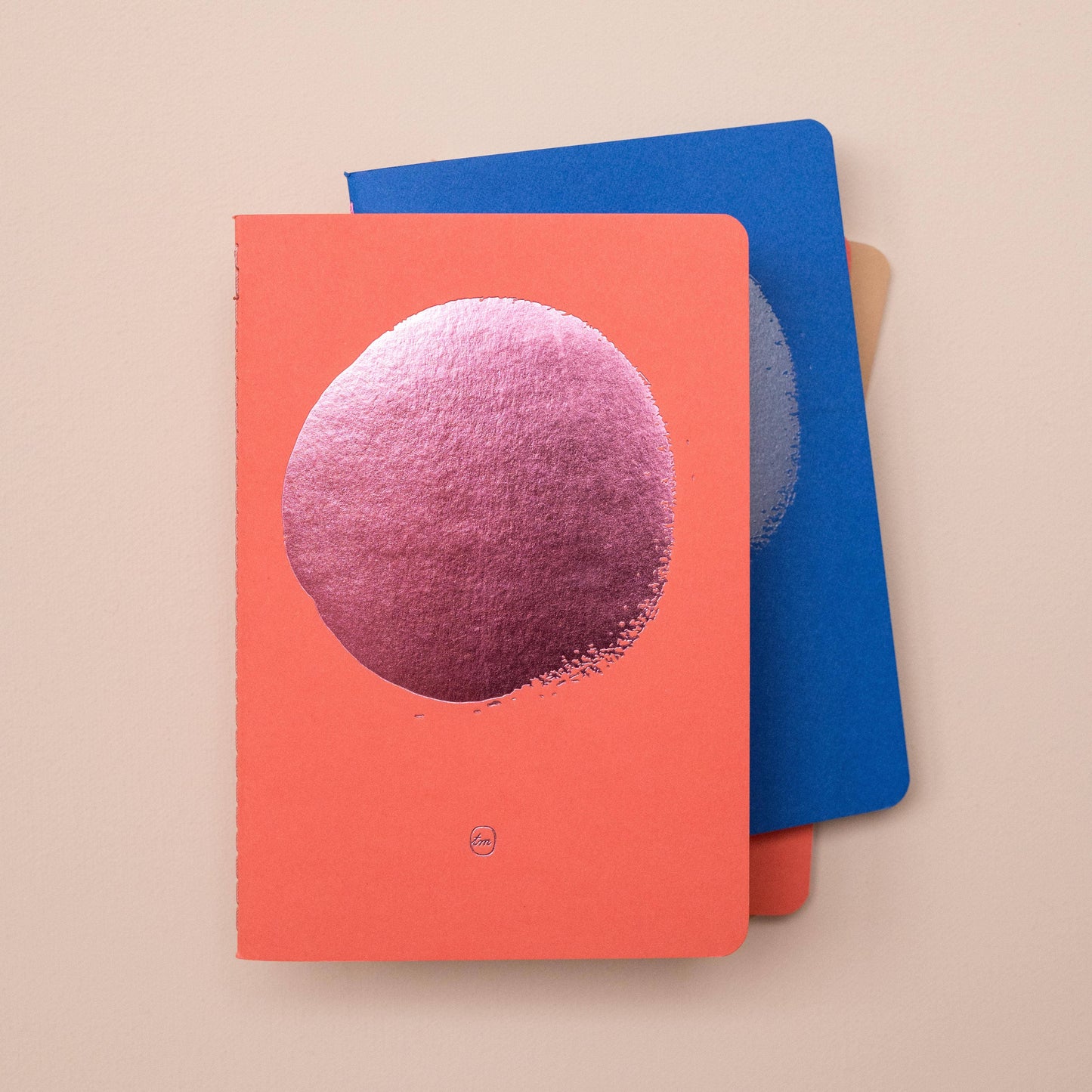 Zwei Tinne Mia Notizbücher ruhen auf einer beigen Oberfläche. Das obere Bullet Journal A6 - Flamingo mit korallenrotem Einband und pinkfarbenem Metallic-Kreis liegt über einem blauen FSC-Mix-Notizbuch, wobei beide den Rand eines braunen Notizbuchs darunter freigeben.