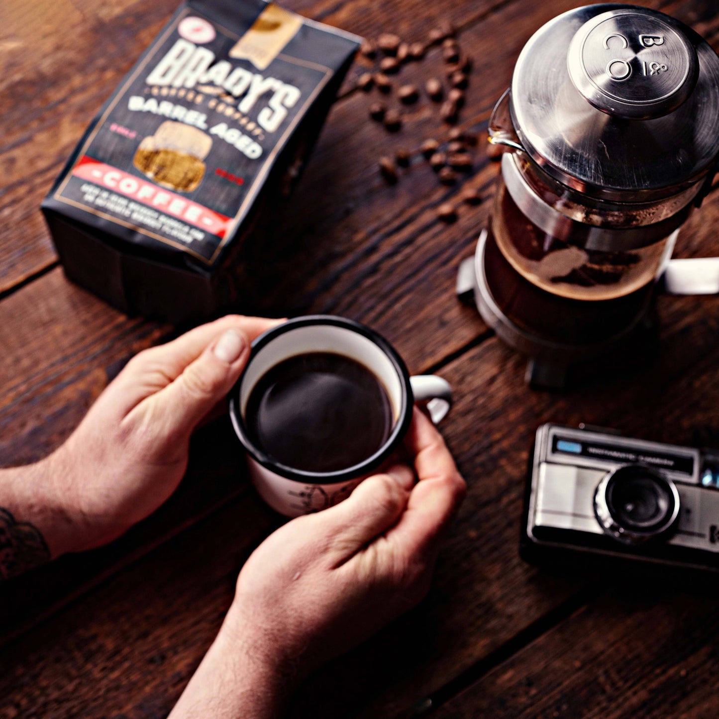Eine Person hält einen Becher mit schwarzem Kaffee auf einem Holztisch mit Brady's Coffee Company - Brady's Barrel Aged Irish Whiskey Kaffeebeutel mit ganzen Bohnen, einer French Press, ganzen Bohnen und einer alten Kamera in der Nähe.