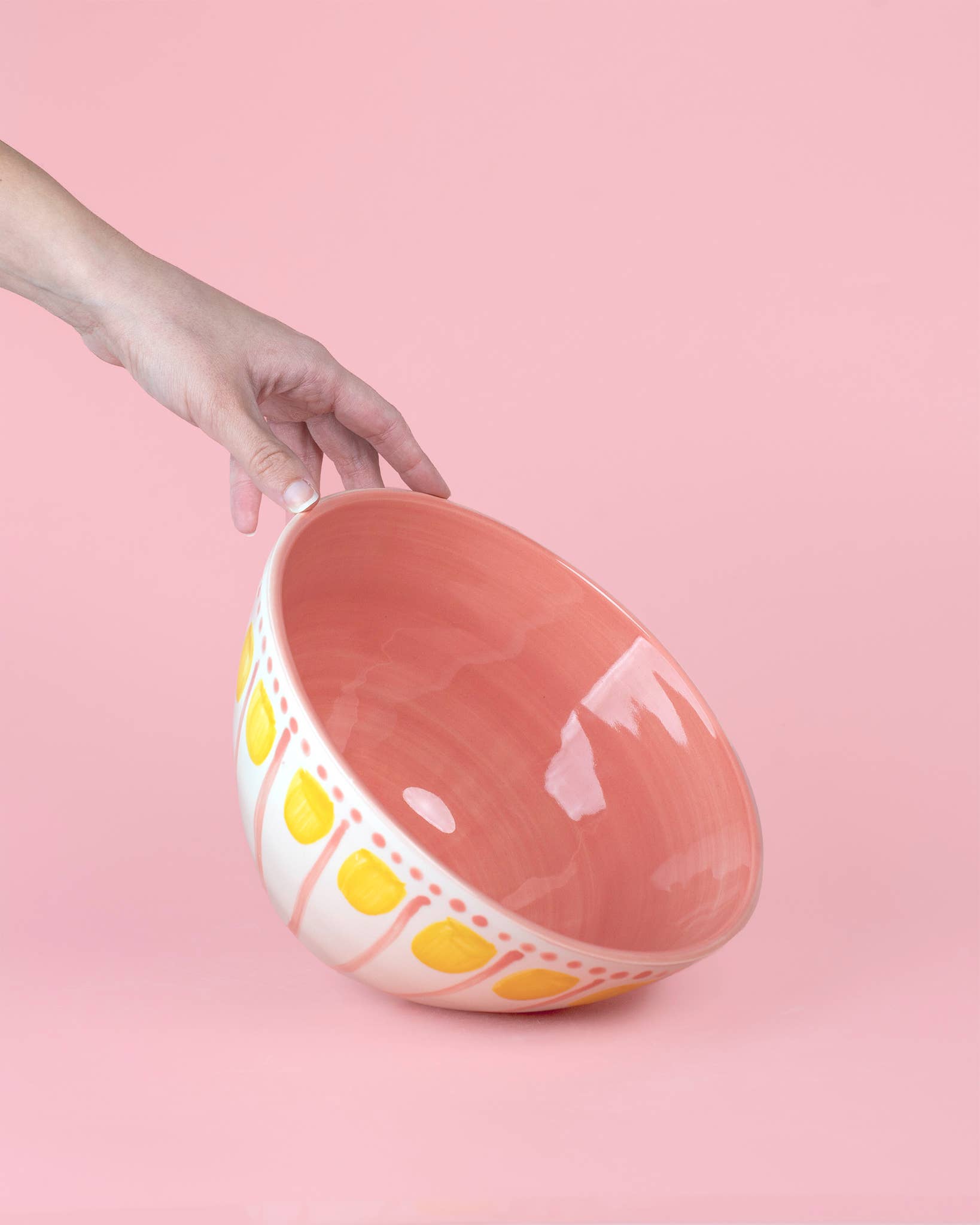 Eine Hand hält die Alfar Tierra Cocida | Handgefertigte Keramik-Servierschale 1,5L Rosa und Gelb VALENTINA, die außen rosa und gelb gemustert und innen rosa ist und spanische Handwerkskunst vor einem unifarbenen rosa Hintergrund präsentiert.