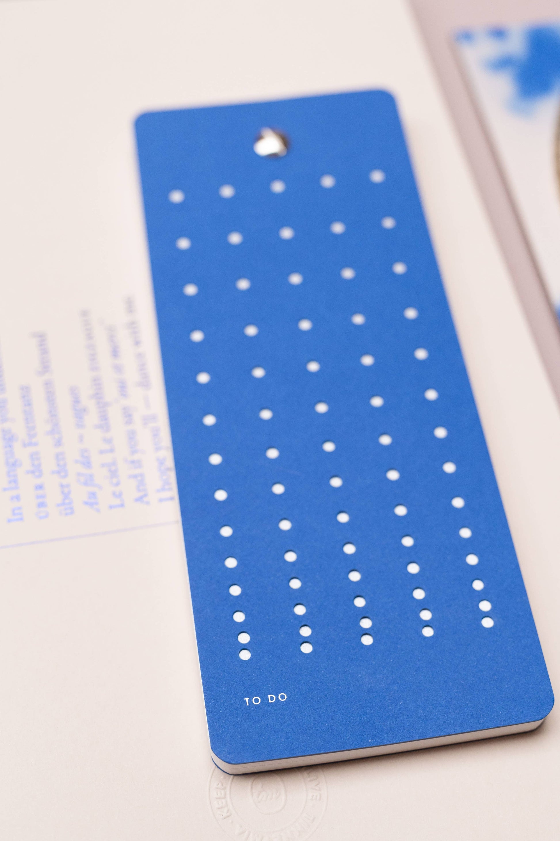 Die Tinne Mia - Aufgabenliste - True Blue ist eine blaue rechteckige Aufgabenliste von Tinne Mia mit gleichmäßig verteilten weißen Punktspalten, die auf einem aufgeschlagenen Buch mit verschwommenem Text und einem schwachen blauen Bild im Hintergrund liegt.